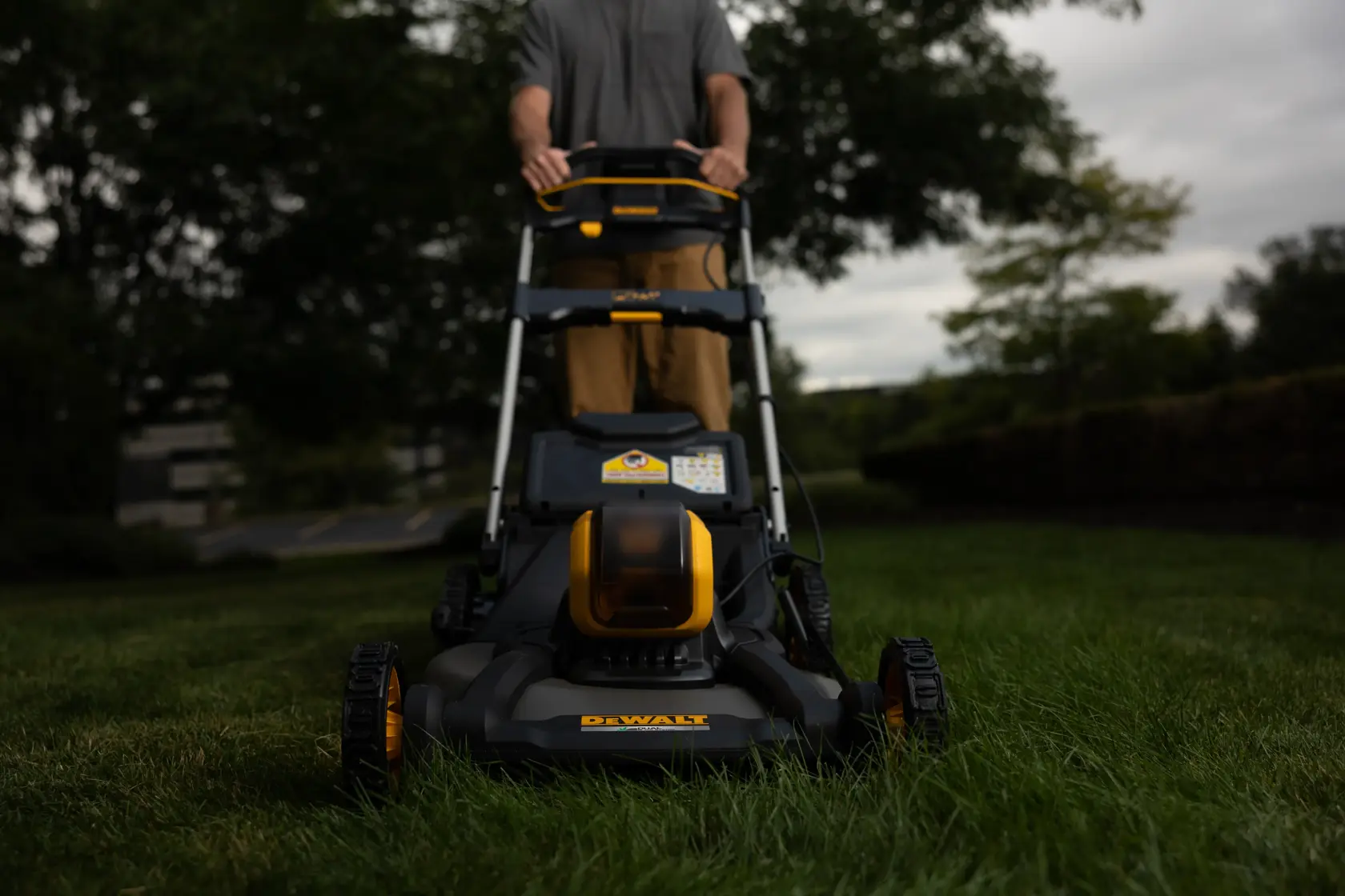 A DEWALT cordless lawn mower is being pushed across a grassy lawn outdoors, with a person standing behind it. The mower has