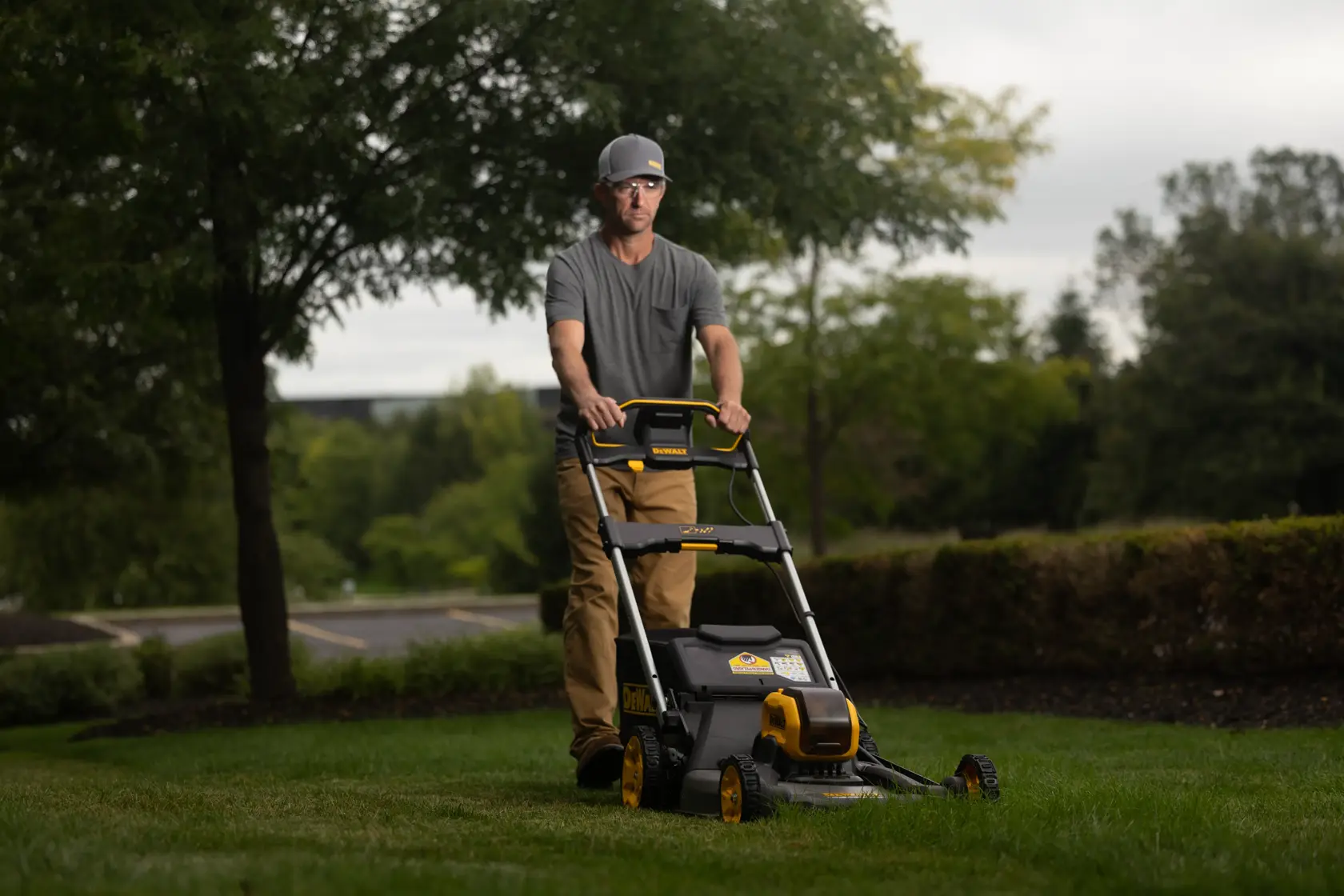 A person with a blurred face is using a DEWALT lawn mower outdoors on a grassy area, surrounded by trees and bushes.