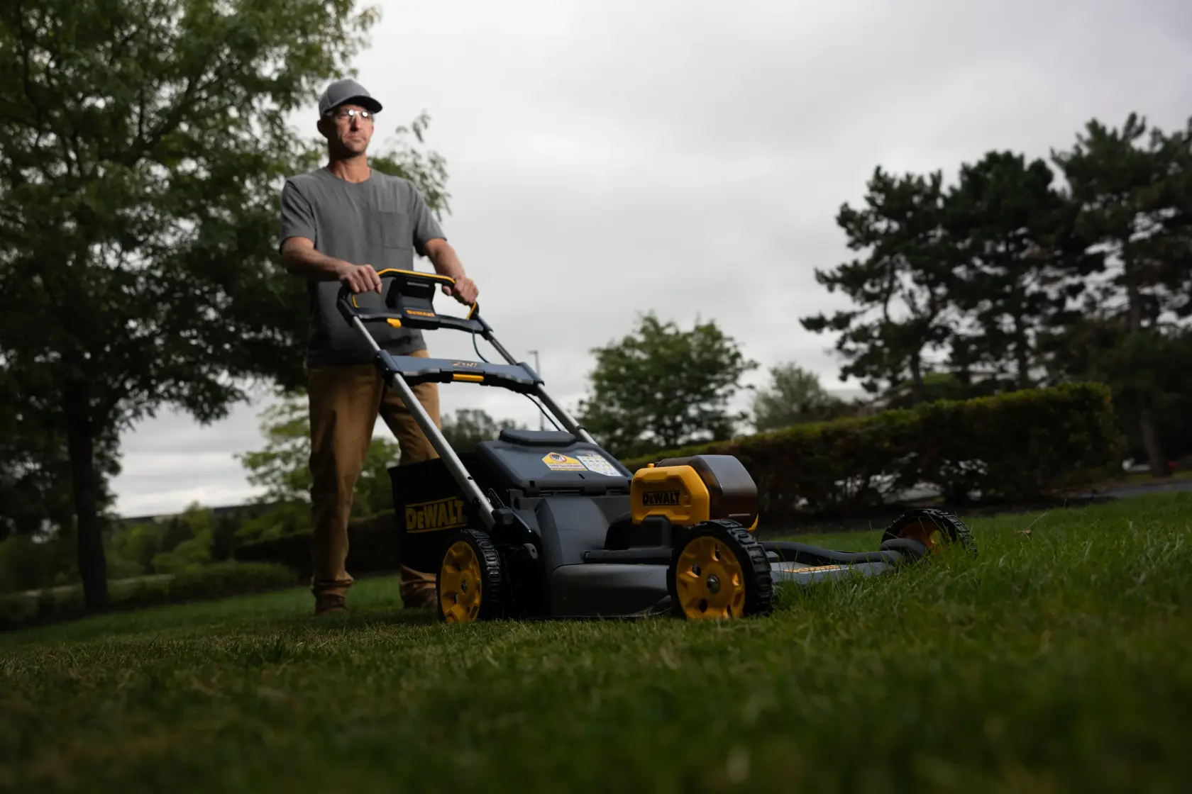 A person uses a DEWALT cordless lawn mower outdoors, surrounded by trees and grass on a cloudy day.