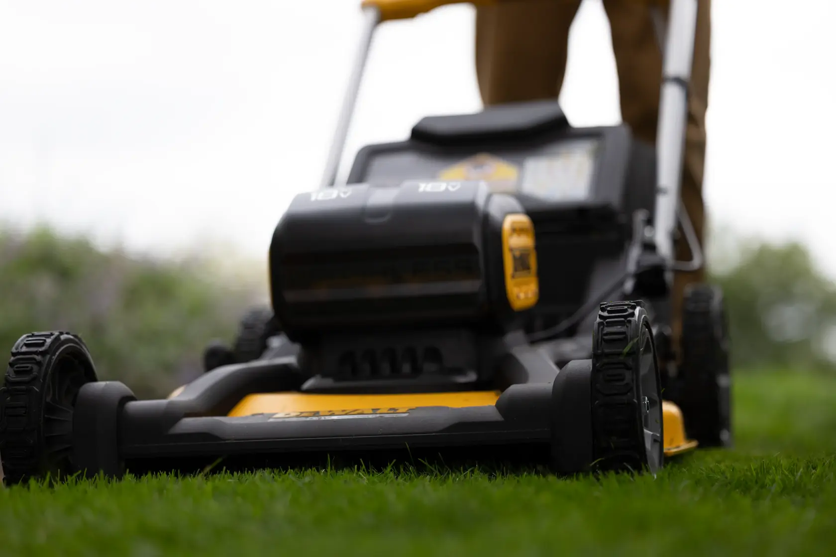 Close-up view of a DEWALT cordless lawn mower being used outdoors on grass, with the lower part of a person visible behind the mower.