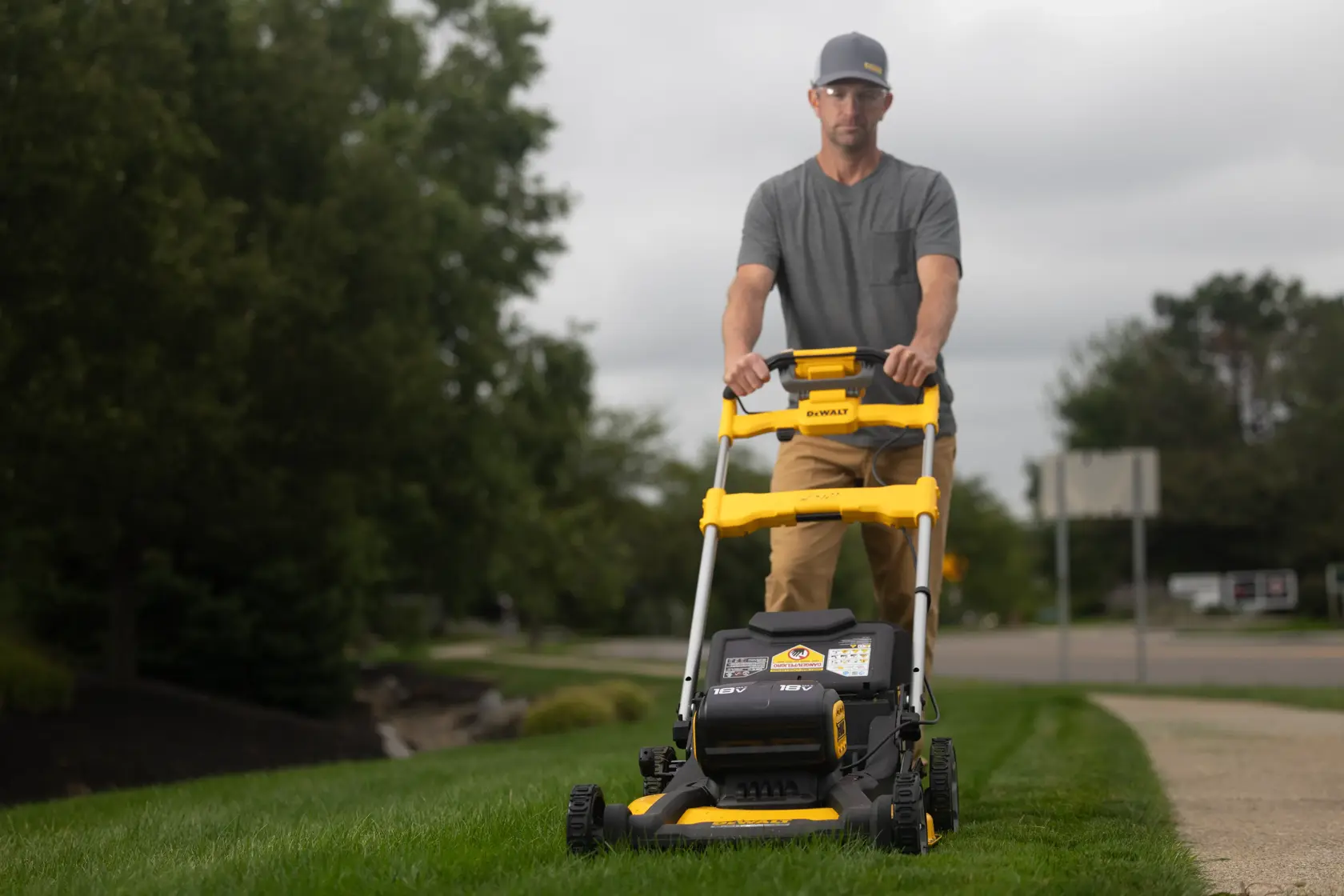 A person is pushing a yellow and black DEWALT lawn mower on a grassy area near a sidewalk. Trees and a street are visible in the background.