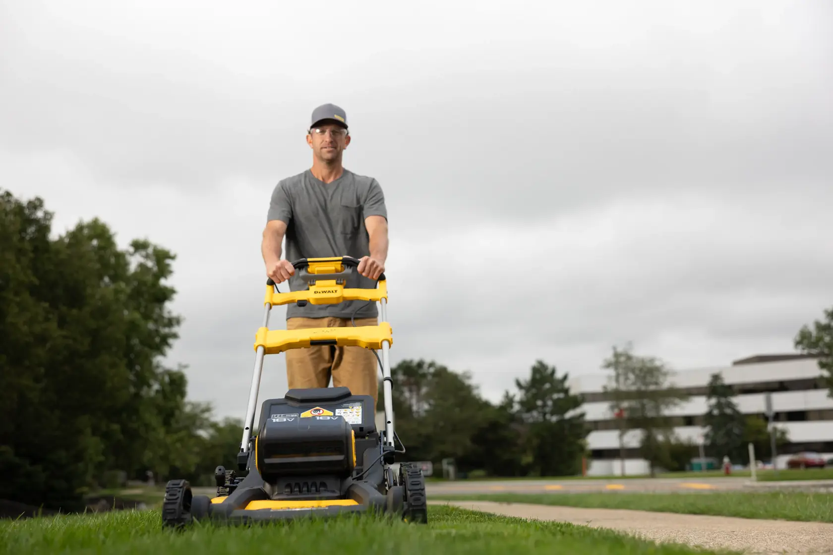 A person is pushing a yellow and black DEWALT lawn mower on a grassy area, with buildings and trees in the background. The person's face is blurred.