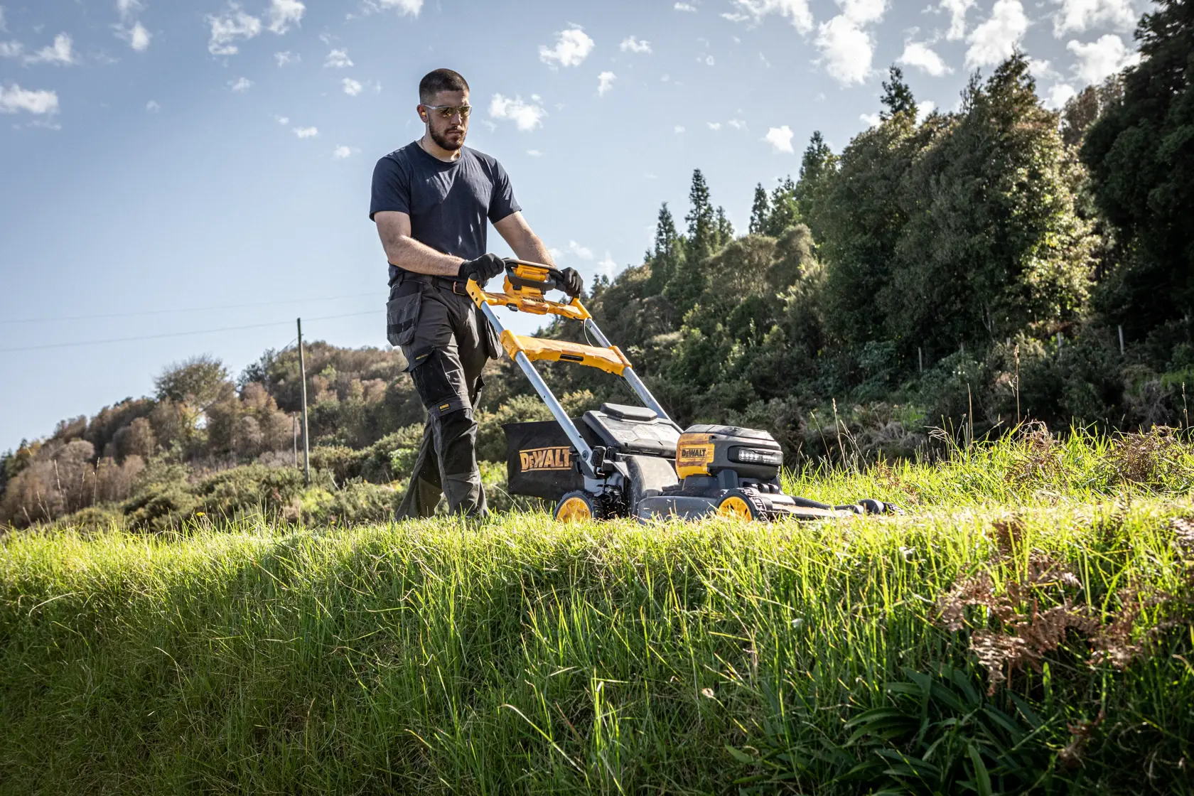 Persona taglia l’erba all’aperto con un tagliaerba DEWALT, giornata soleggiata e alberi sullo sfondo.