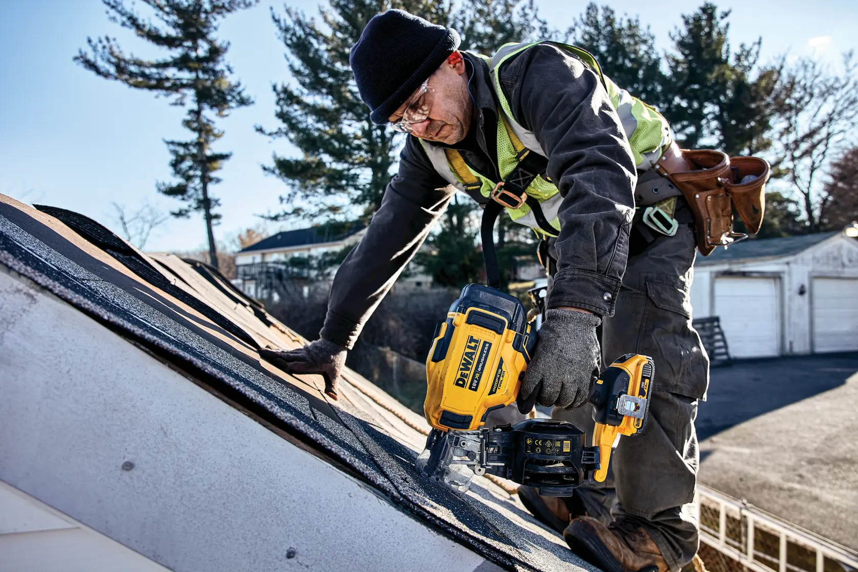 Worker uses DEWALT roofing nailer on a shingled roof outdoors. Safety gear and toolbelt visible.
