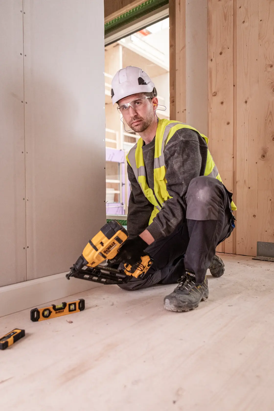 Worker uses DEWALT DCN660N nailer on wooden construction floor, wearing safety gear and vest.