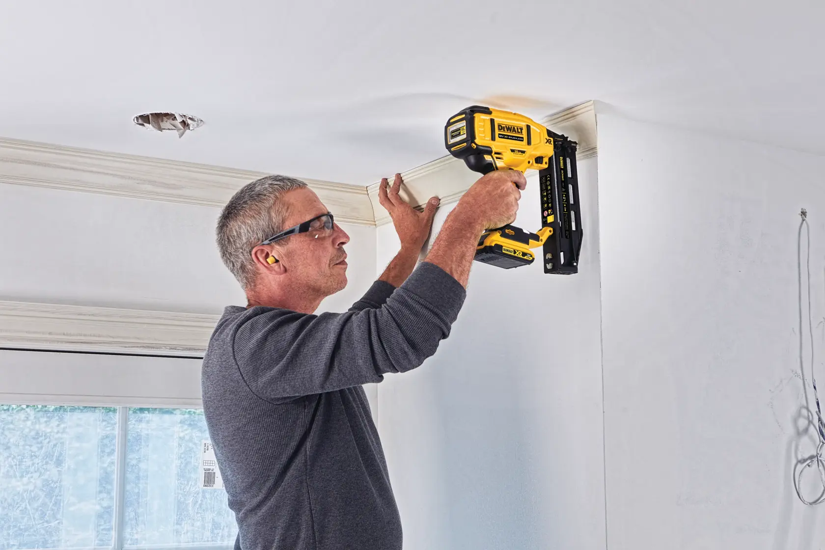 Person uses DEWALT DCN662N nailer to secure crown molding to a ceiling in a bright room.