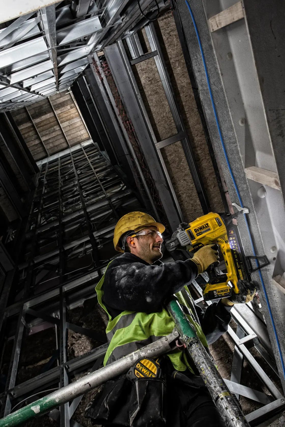 Worker using DEWALT DCN890P2 nailer on metal framework, wearing safety gear in construction site.