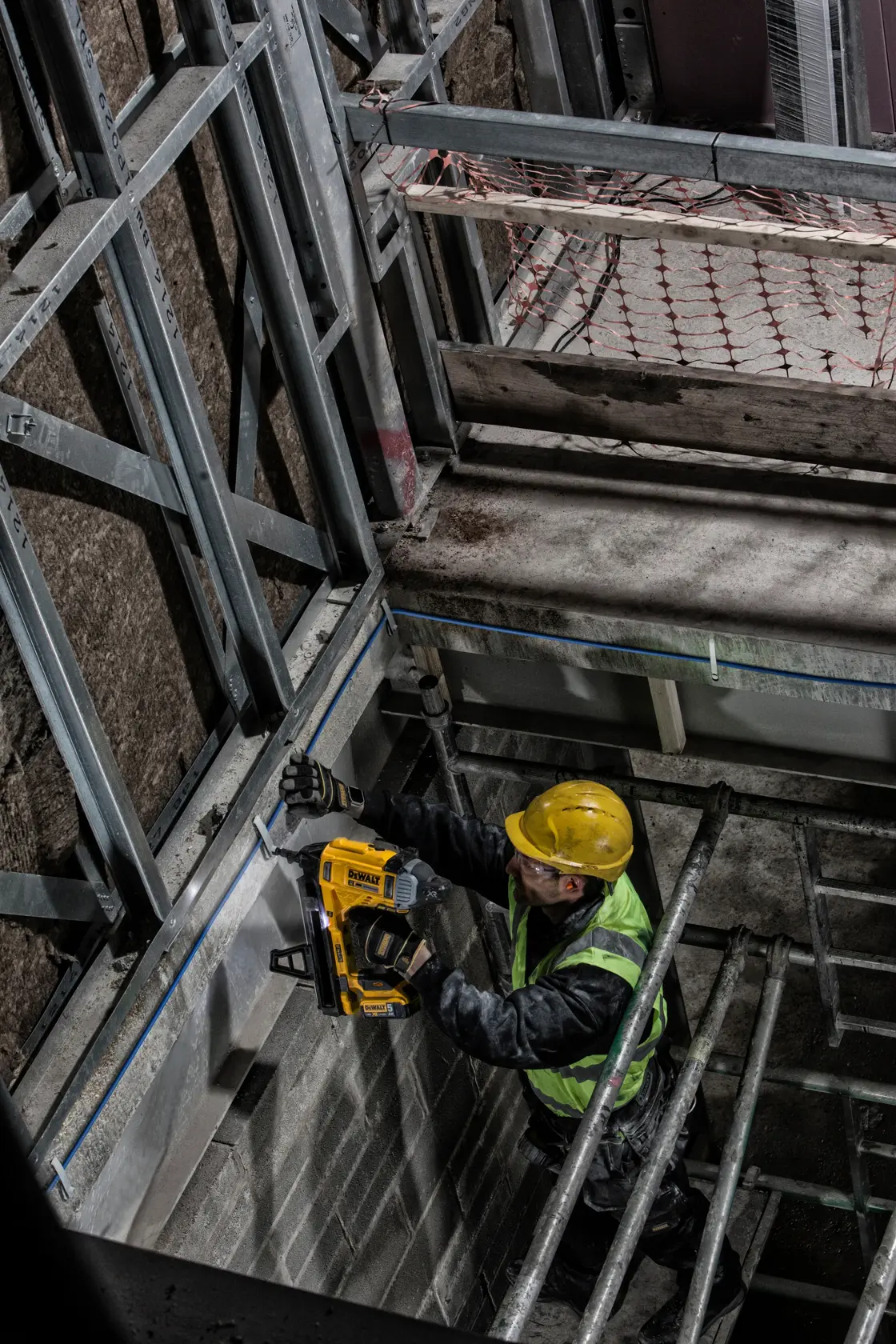 Worker uses DEWALT DCN890P2 nailer on wall frame at a construction site, wearing safety gear.