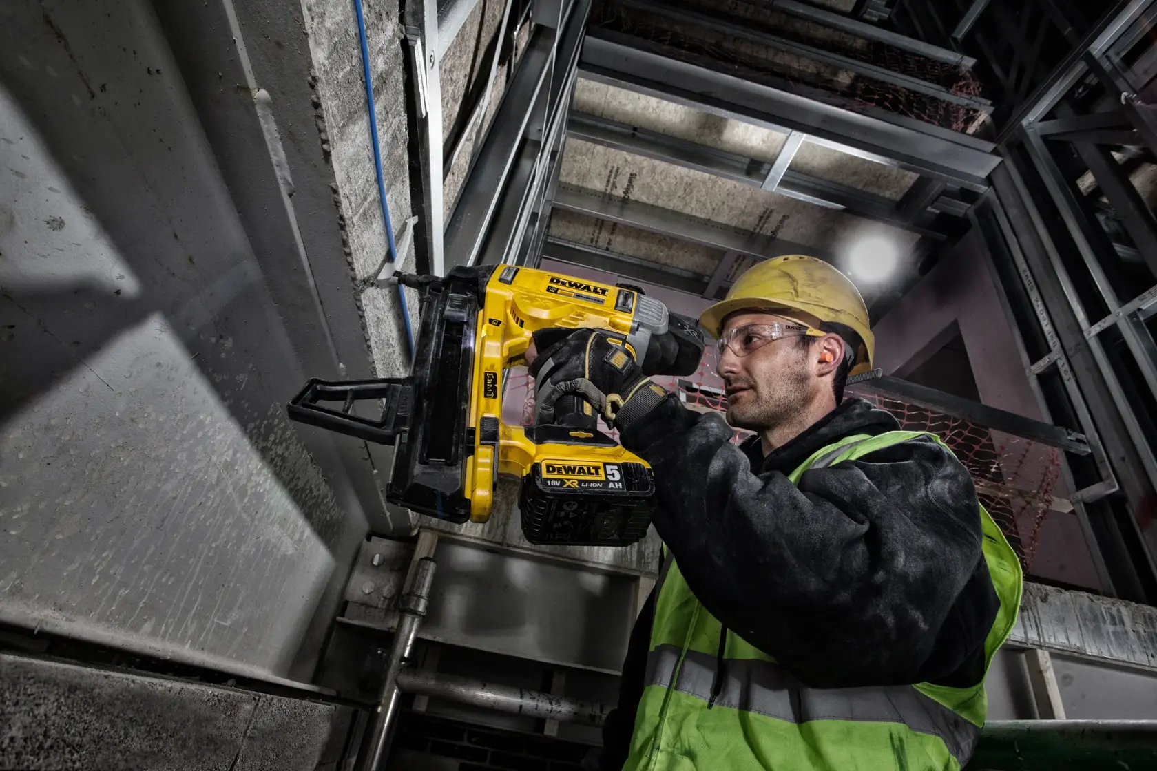 Worker uses DEWALT DCN890P2 nailer on metal frame in industrial setting, wearing safety gear and helmet.