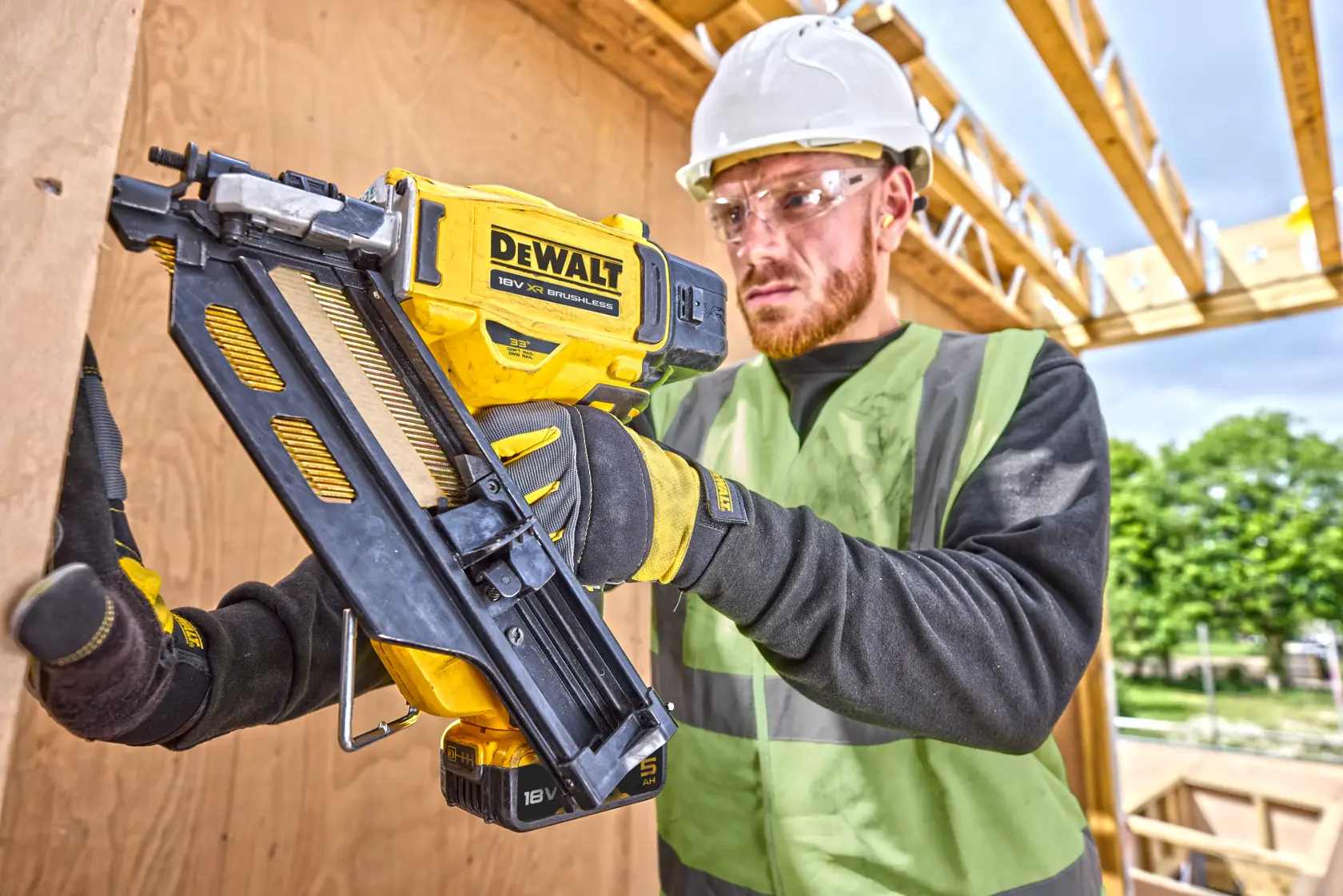 Worker uses DEWALT DCN950N nailer on wood at construction site, wearing safety gear and gloves.