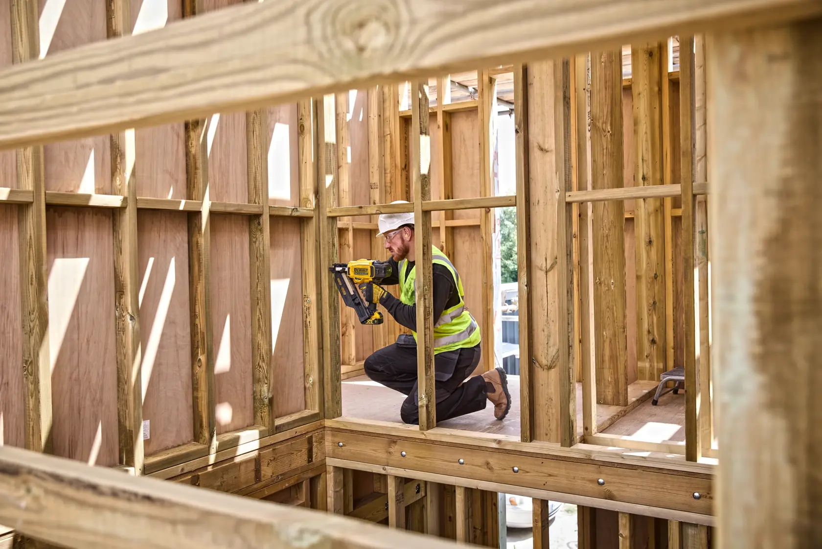 Person using DEWALT tool on wooden frame at construction site, wearing safety vest and kneeling indoors.