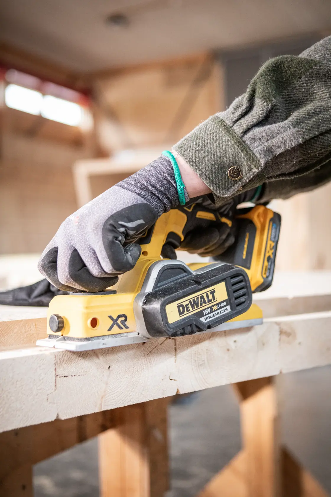 Gloved hand using a DEWALT cordless planer on wood in a workshop setting.