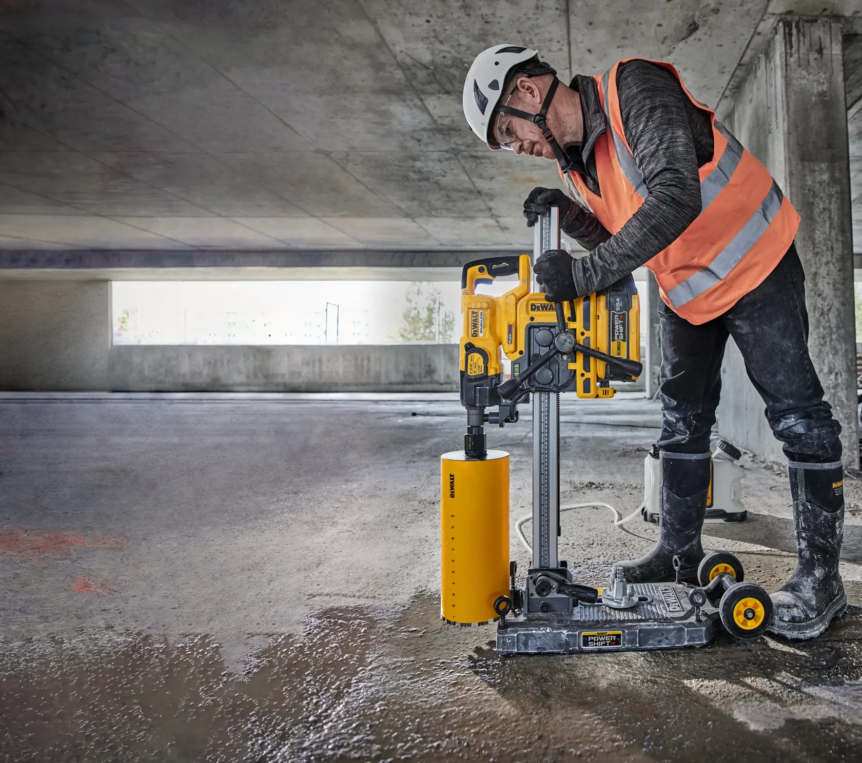 Worker uses DEWALT DCPS151 core drill on wet concrete in a parking structure, wearing safety gear.