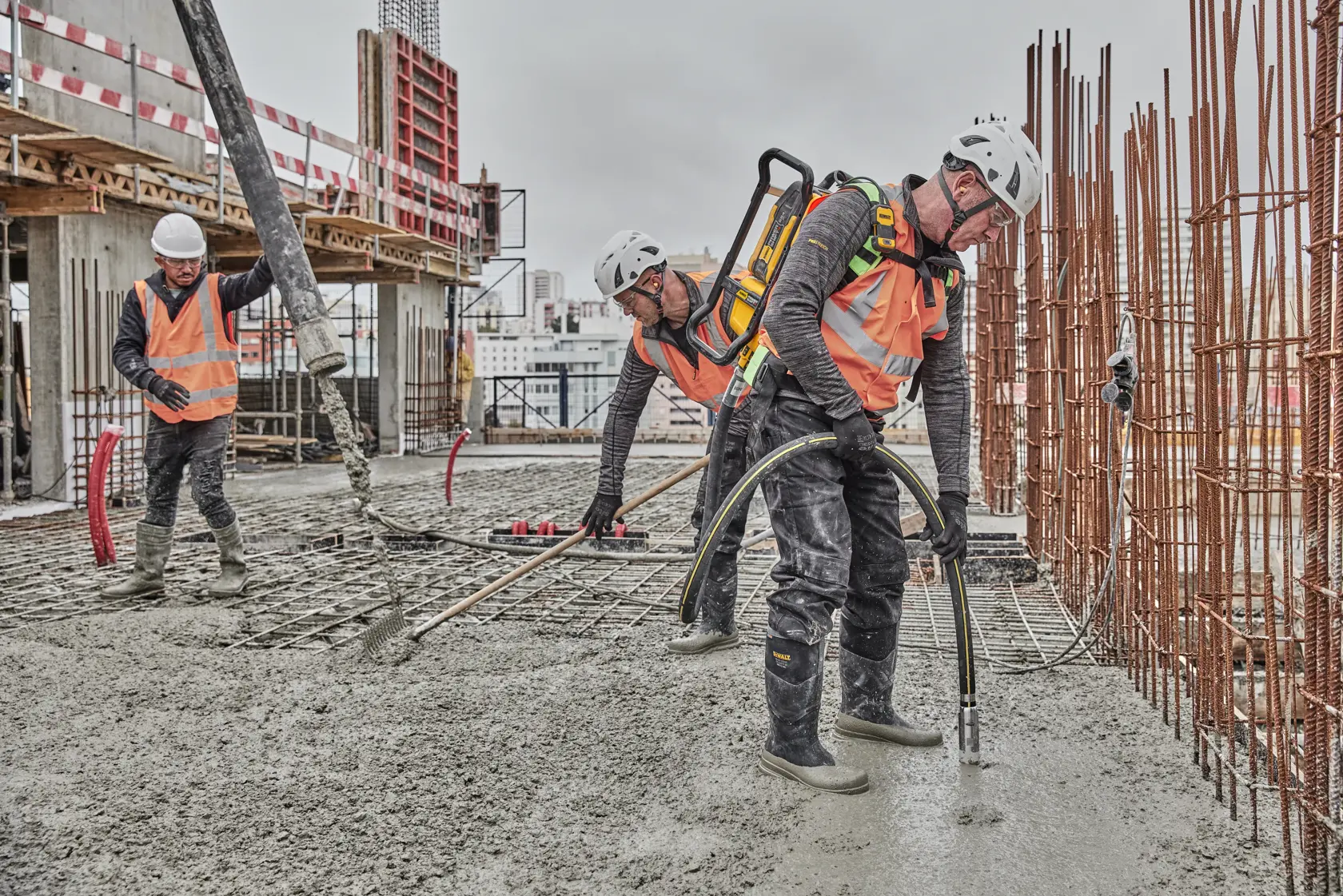 Construction workers in safety gear operate concrete equipment on a building site with rebar and wet cement.
