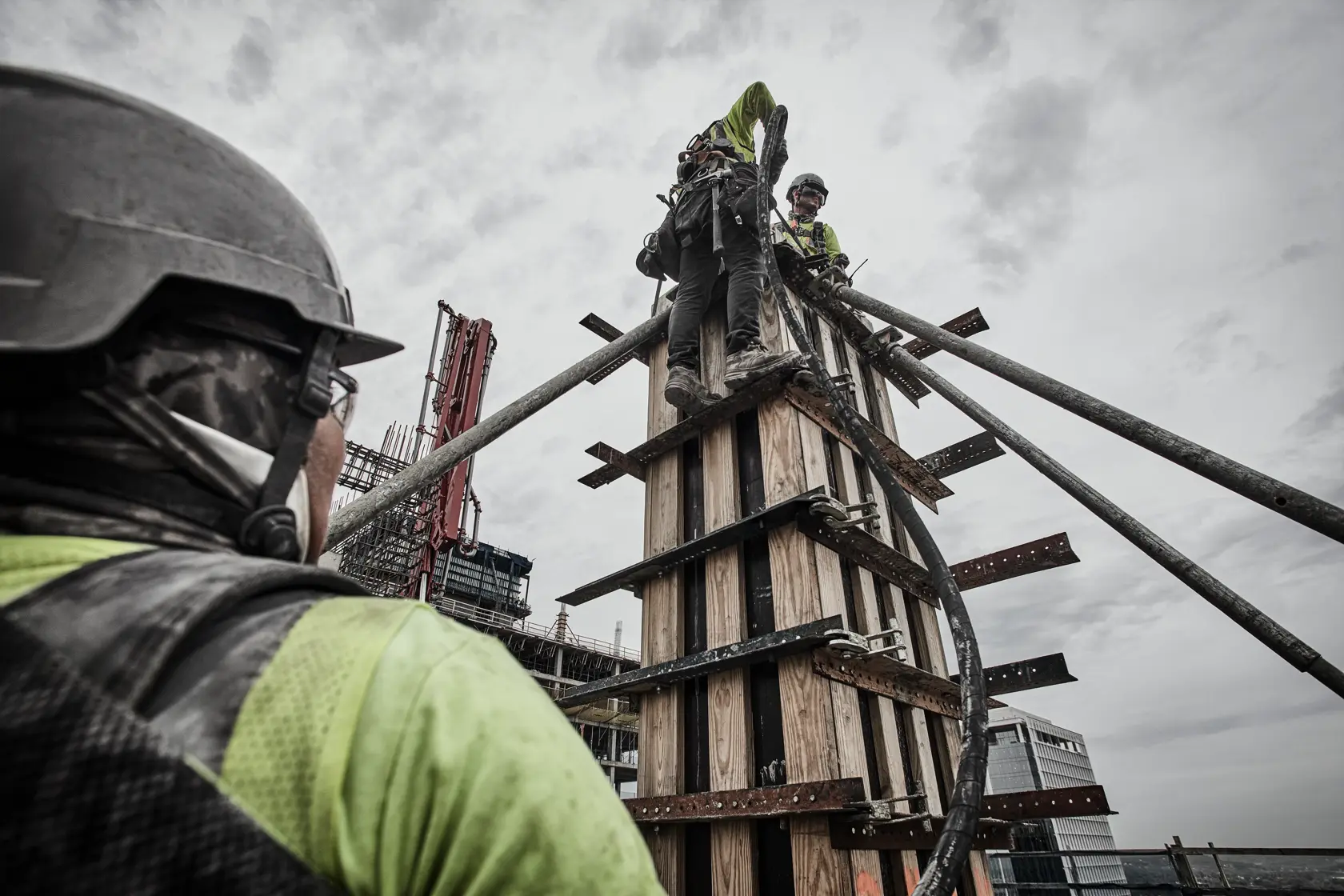 Workers in safety gear on a construction site, assembling wooden framework with pipes, under a cloudy sky.