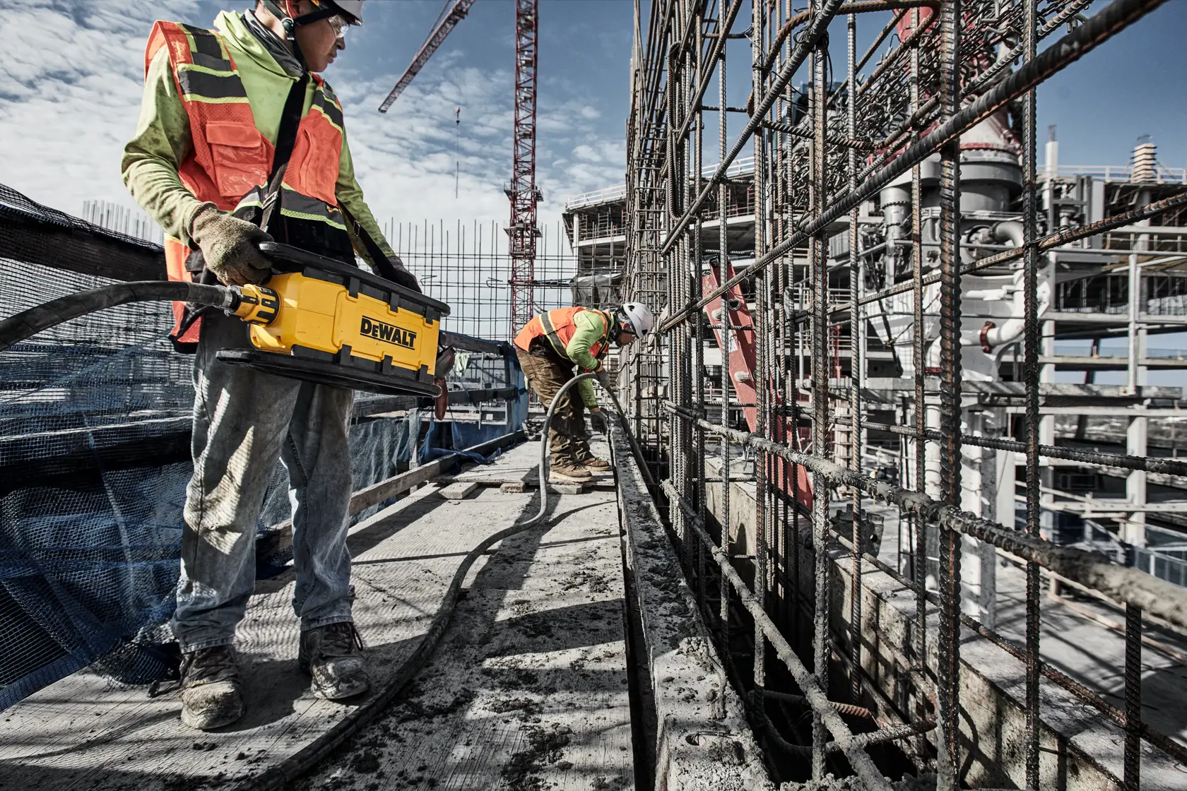 Two workers use a DEWALT tool at a construction site, surrounded by steel bars and scaffolding.