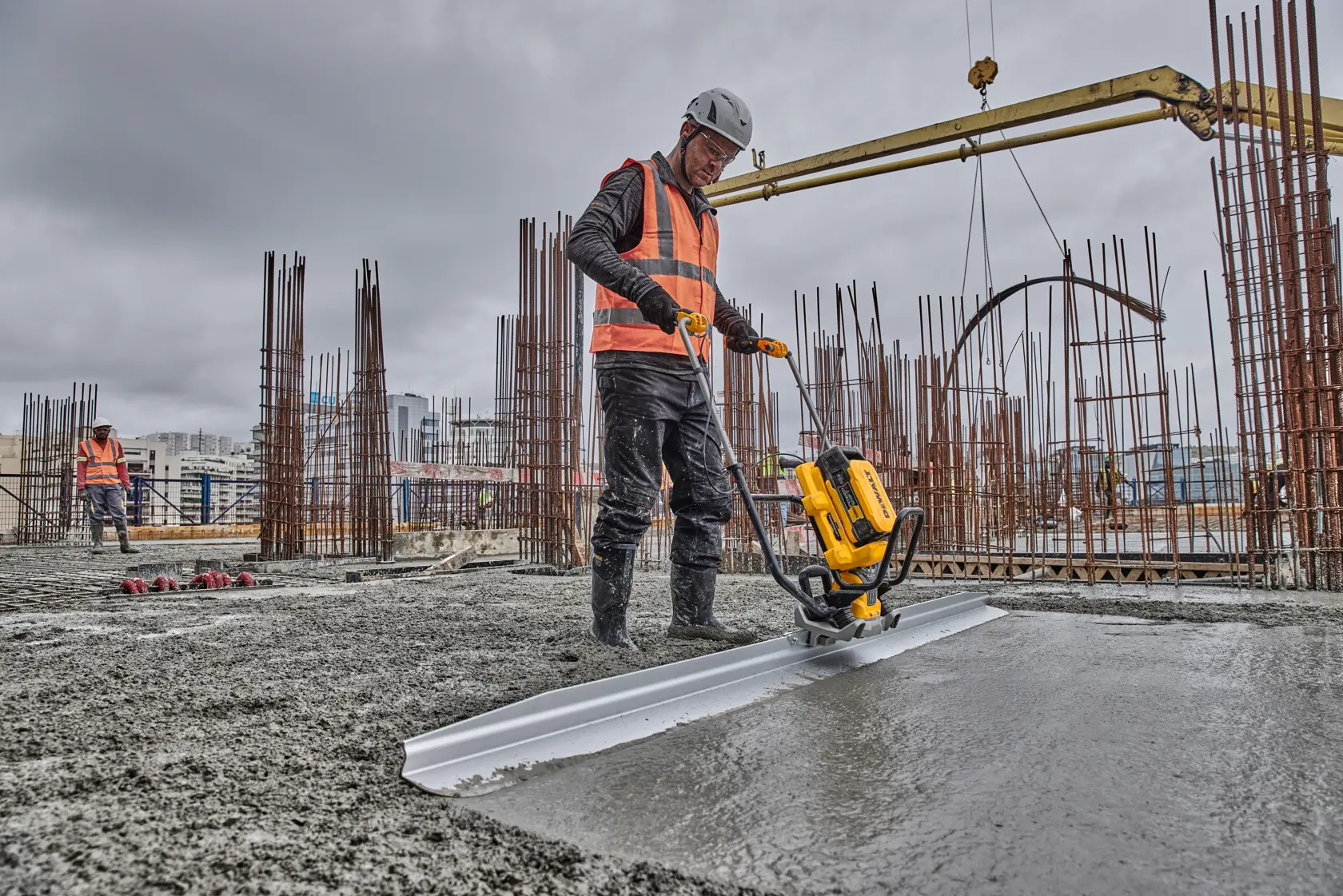 Worker uses DEWALT screed to smooth wet concrete at a construction site with steel rods and machinery visible.
