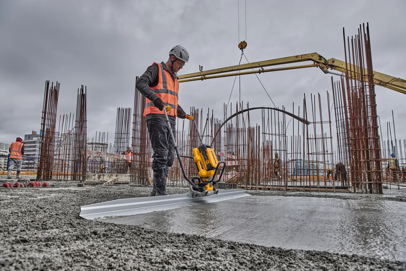 Worker uses DEWALT tool to level concrete on a construction site with steel rods and heavy machinery.
