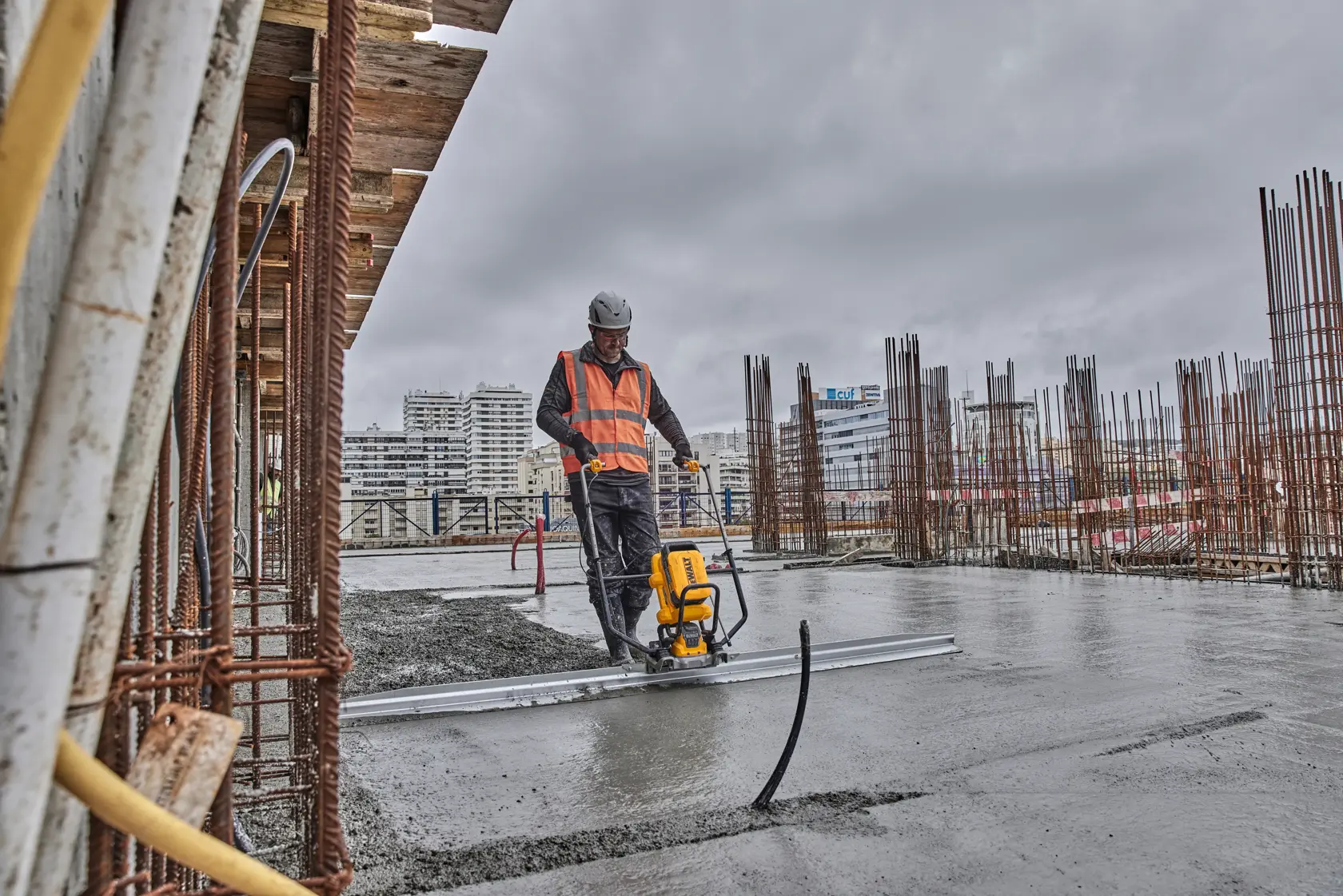 Worker using DEWALT tool to smooth concrete on construction site with buildings and steel rods in background.