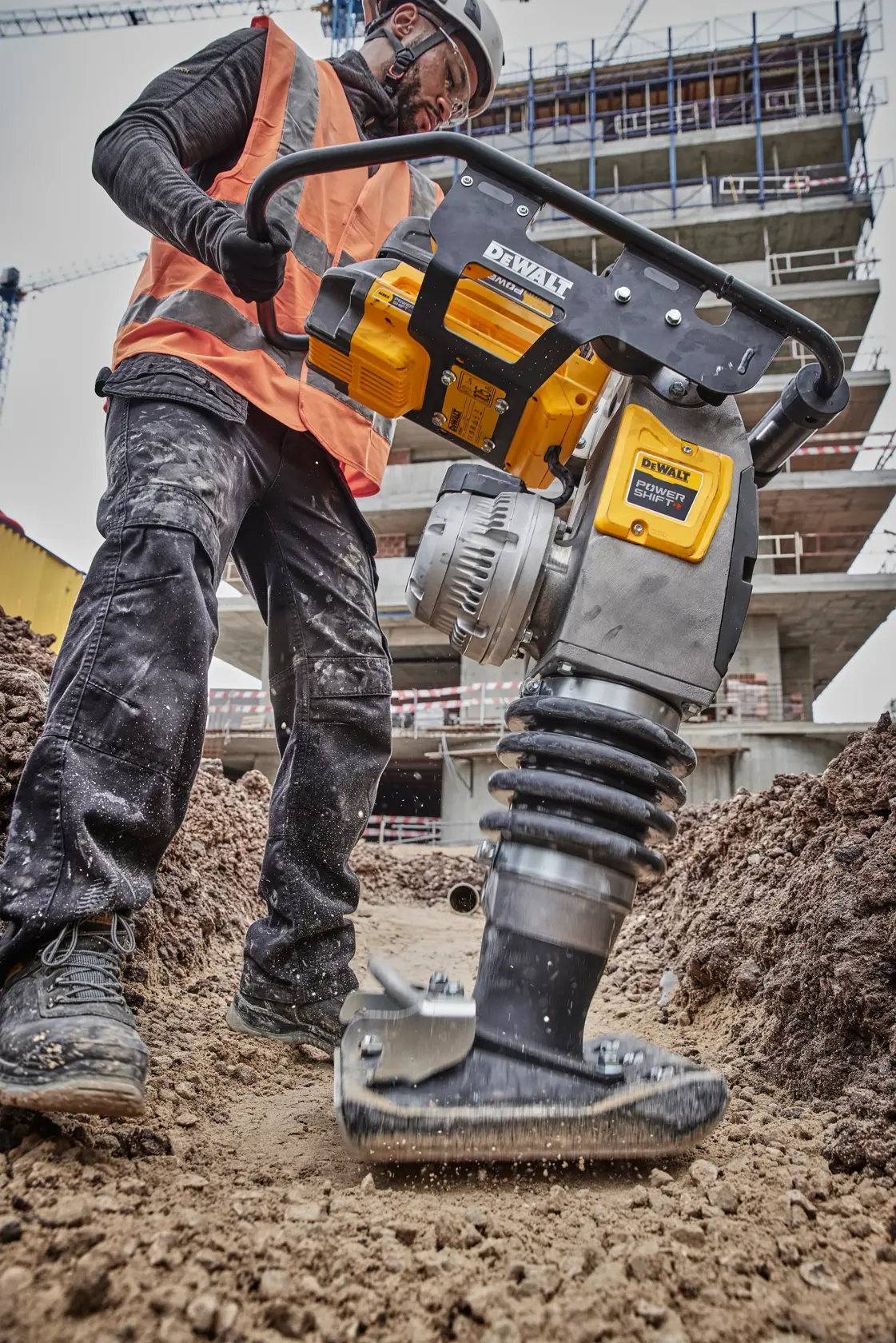Worker uses DEWALT compactor DCPS660N on soil at construction site with buildings and scaffolding in the background.