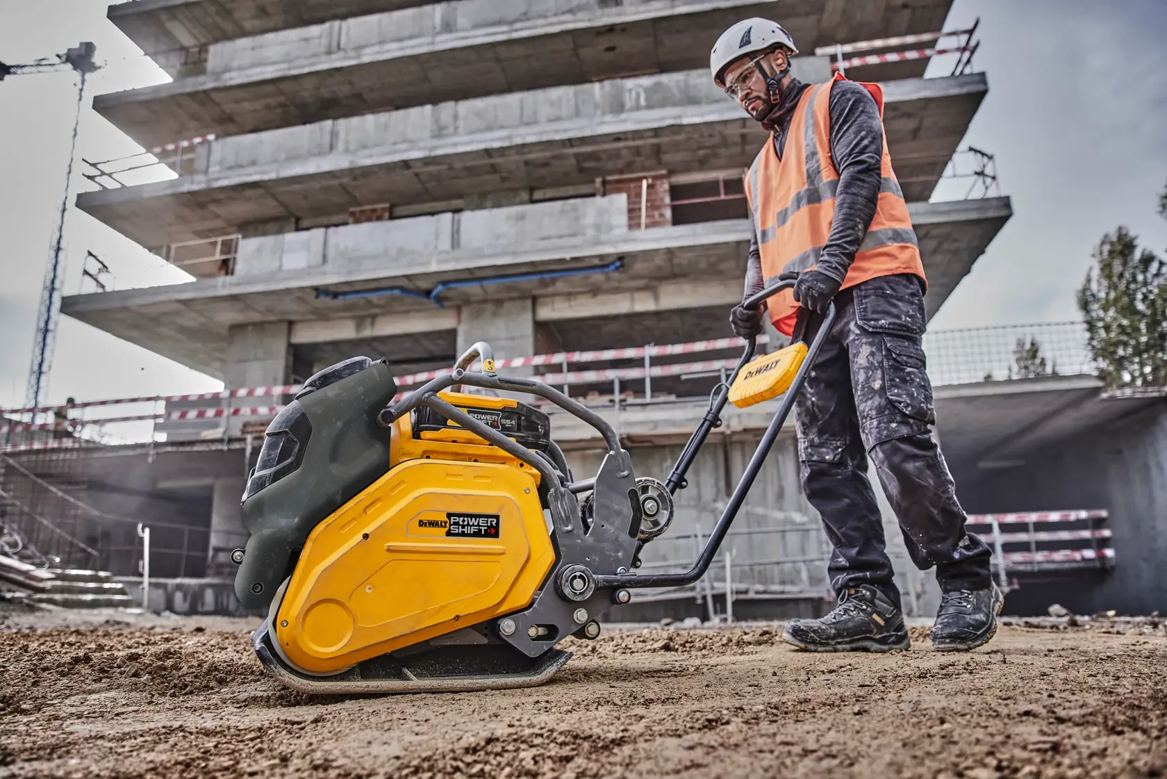 Worker uses DEWALT plate compactor at construction site, wearing safety gear and vest.