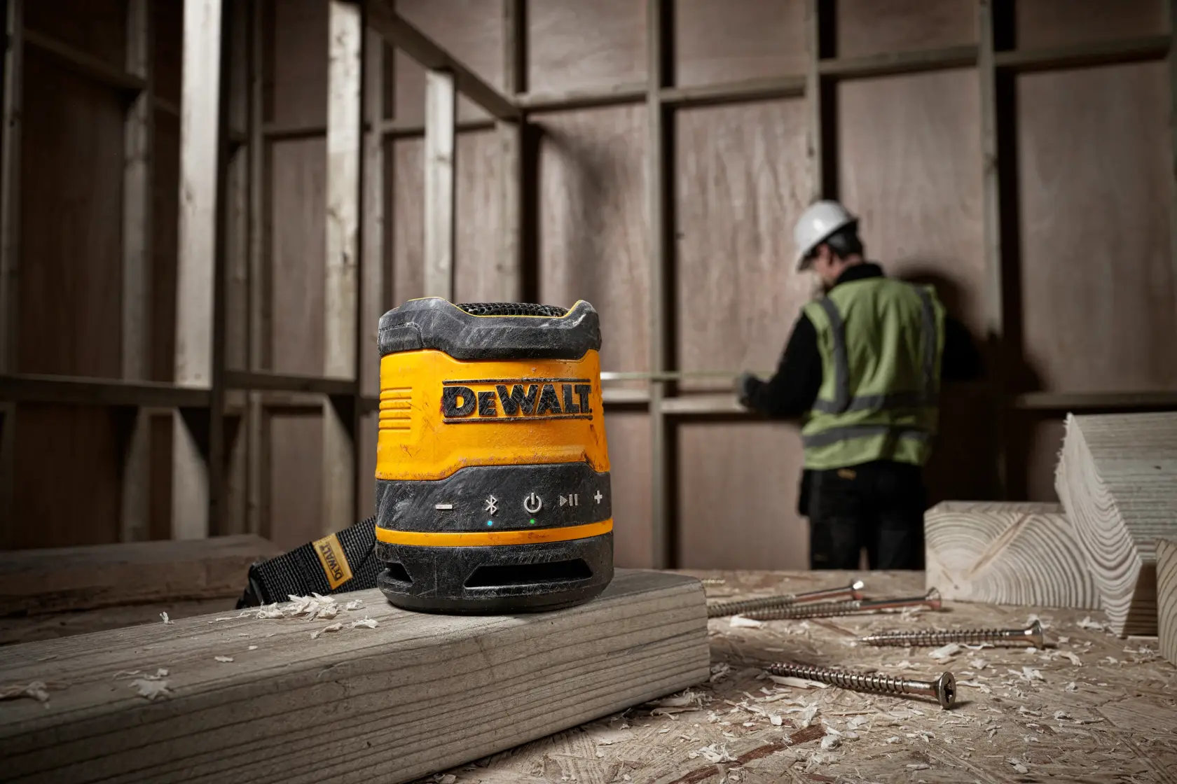 En bærbar Bluetooth-høyttaler DEWALT DCR009 plassert på en treplanke på en byggeplass, med en arbeider i sikkerhetsvest og hjelm som måler i bakgrunnen.