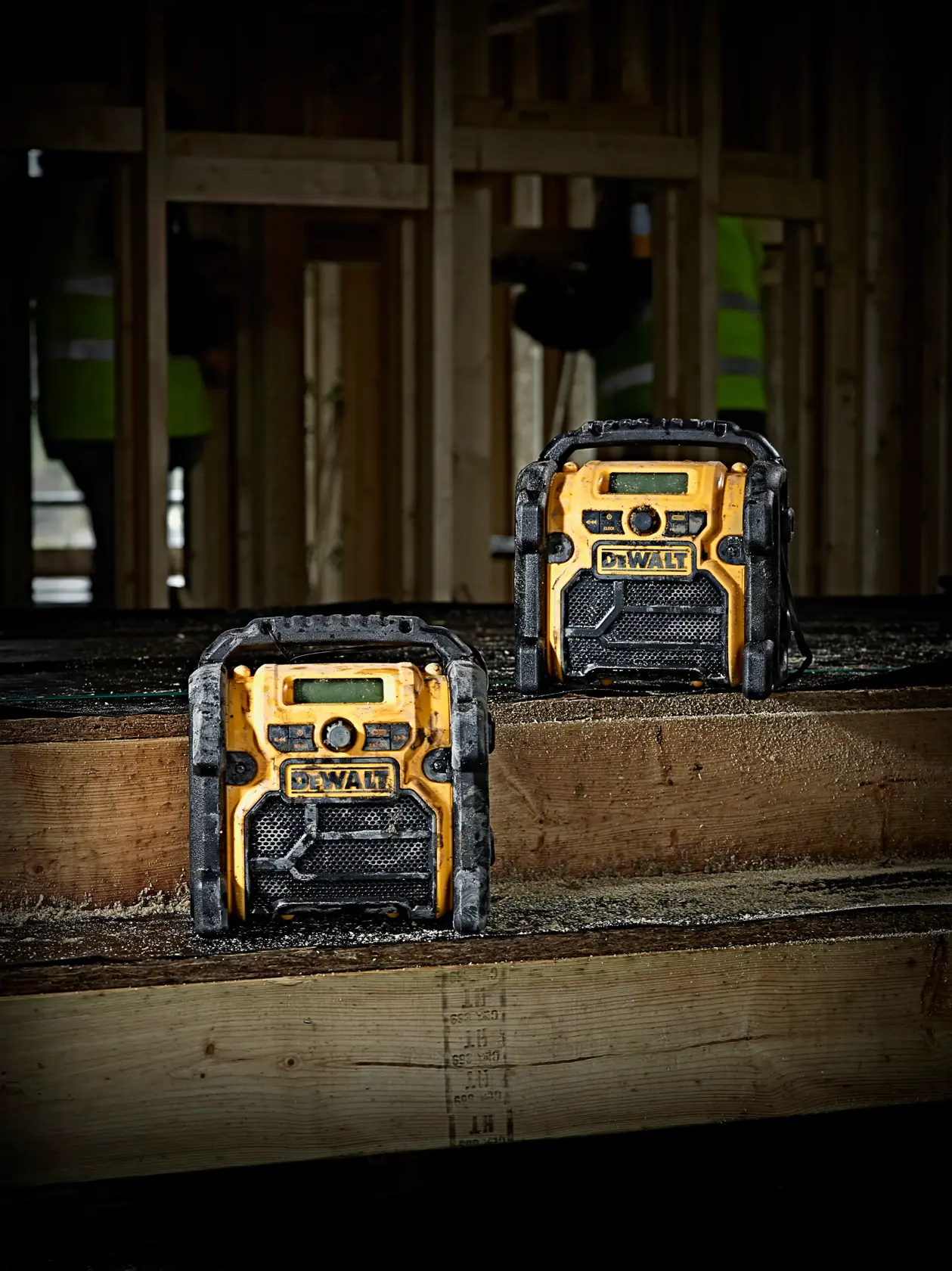 Two DEWALT DCR019 radios on a wooden workbench in a construction site with unfinished wood framing.