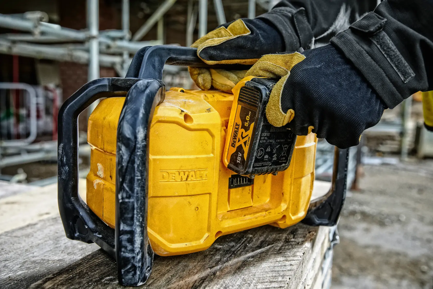 Gloved hands insert battery into yellow DEWALT device at construction site with scaffolding in background.