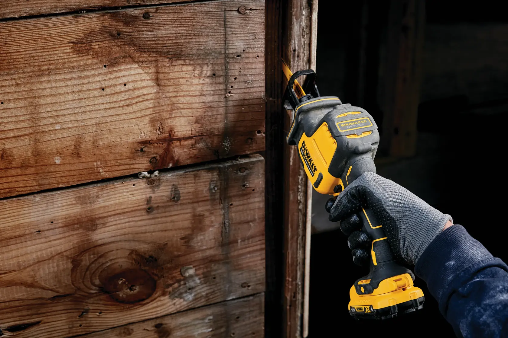 Gloved hand using a DEWALT cordless reciprocating saw on a wooden surface indoors.