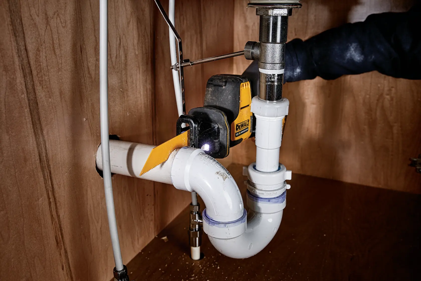 DEWALT cordless saw cutting a PVC pipe under a sink in a wooden cabinet.