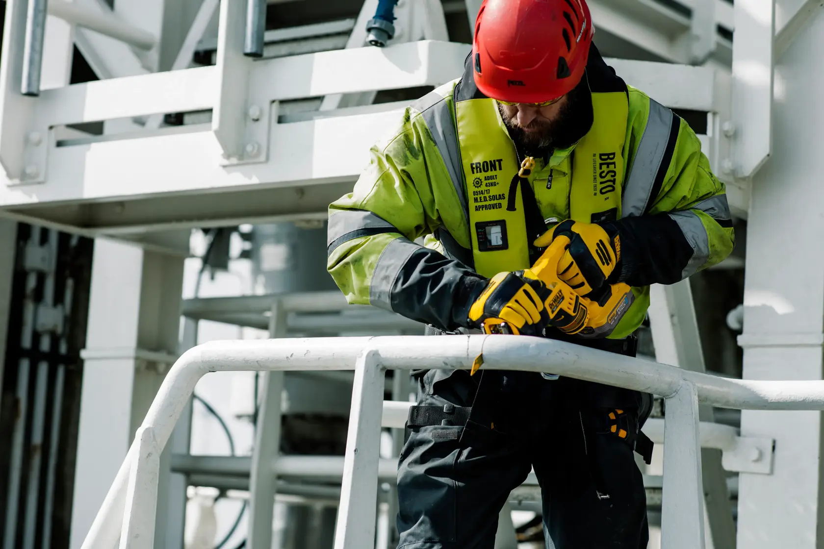 En person med rød vernehjelm og refleksjakke bruker et gult DEWALT verktøy for å kutte metallrekkverk på en industriarbeidsplass.