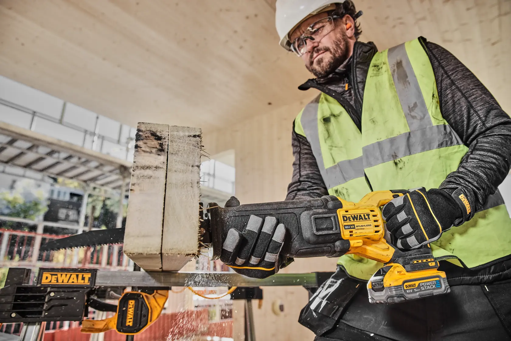 En bygningsarbeider med verneutstyr, inkludert hjelm, hansker og synlighetsvest, bruker en DEWALT batteridrevet bajonettsag (modell DCS386H2T) for å kutte gjennom et tykt trestykke på en byggeplass.