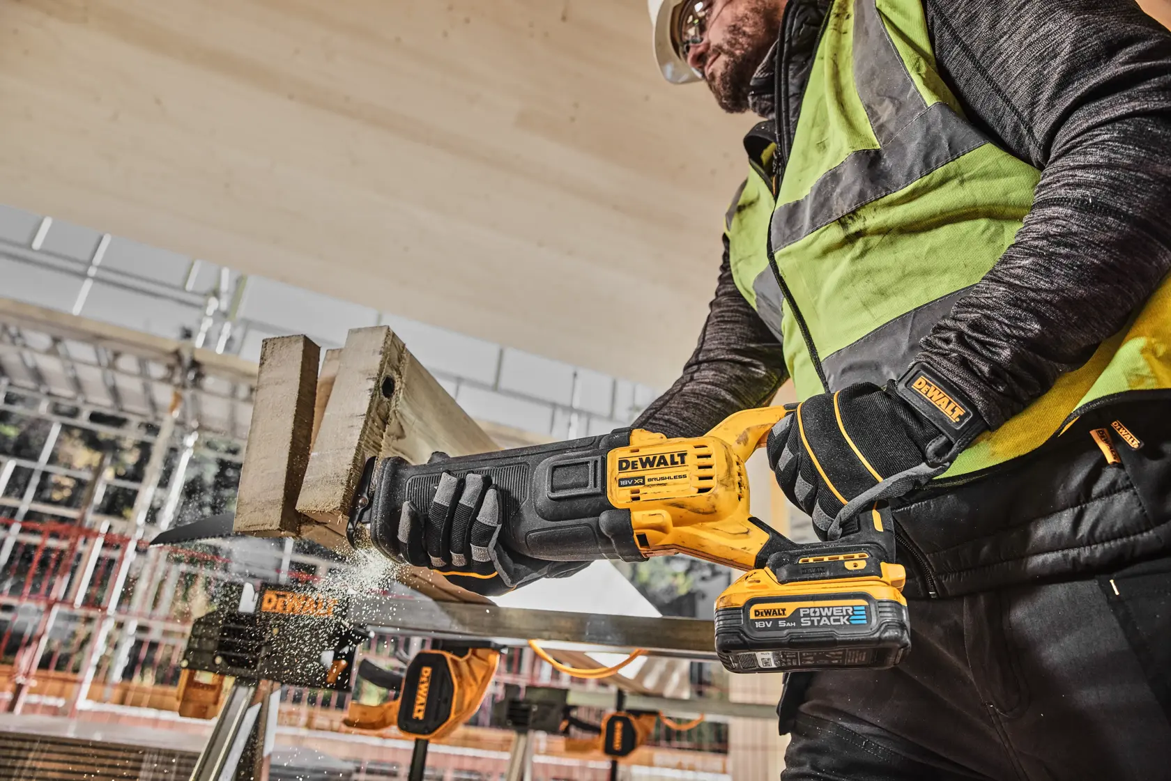 En bygningsarbeider bruker en DEWALT DCS386H2T batteridrevet bajonettsag for å kutte tre på en byggeplass, iført verneutstyr som hansker, vest og hjelm.