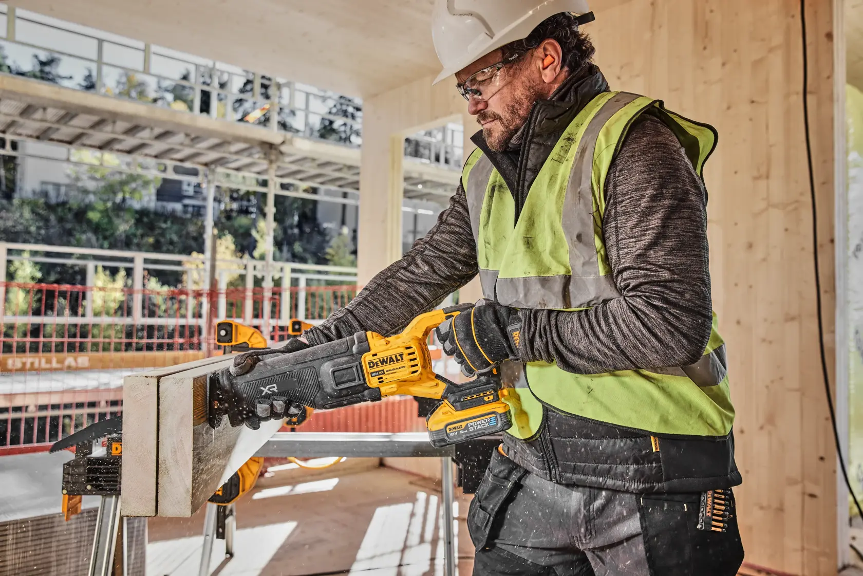 En person med refleksvest og hjelm bruker en DEWALT batteridrevet bajonettsag (modell DCS386H2T) til å kutte en trebit på en byggeplass.