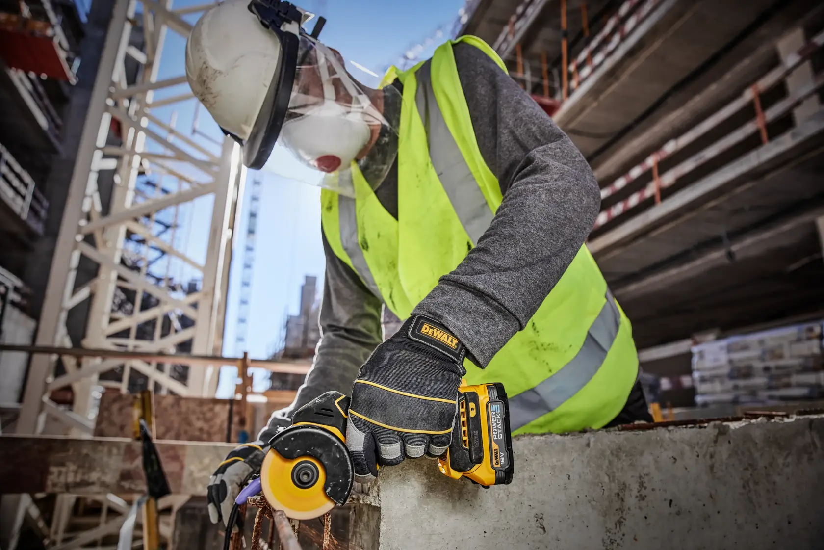 En bygningsarbeider med vernehjelm, ansiktsvisir, refleksvest og hansker bruker et DEWALT DCS438N trådløst verktøy for å skjære i betong på en utendørs byggeplass.