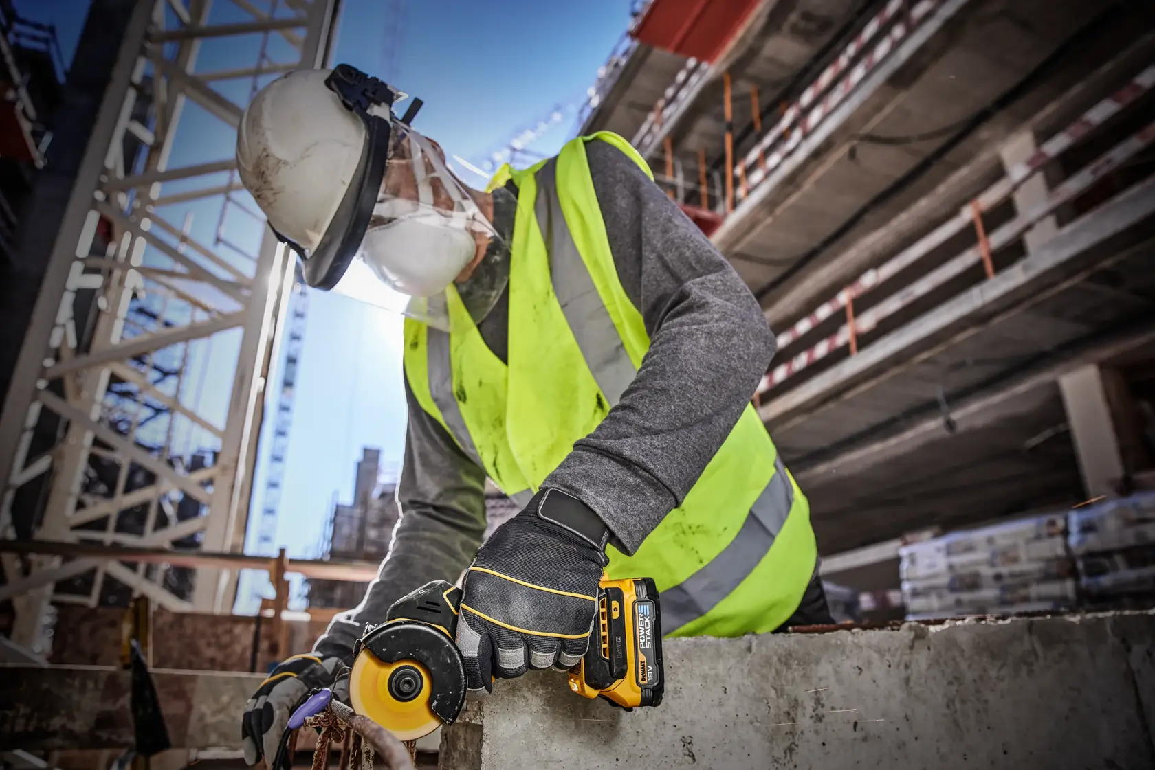 En bygningsarbeider med sikkerhetshjelm, synlighetsvest, hansker og ansiktsskjerm bruker et DEWALT DCS438N batteridrevet verktøy for å kutte eller slipe betong på en byggeplass.