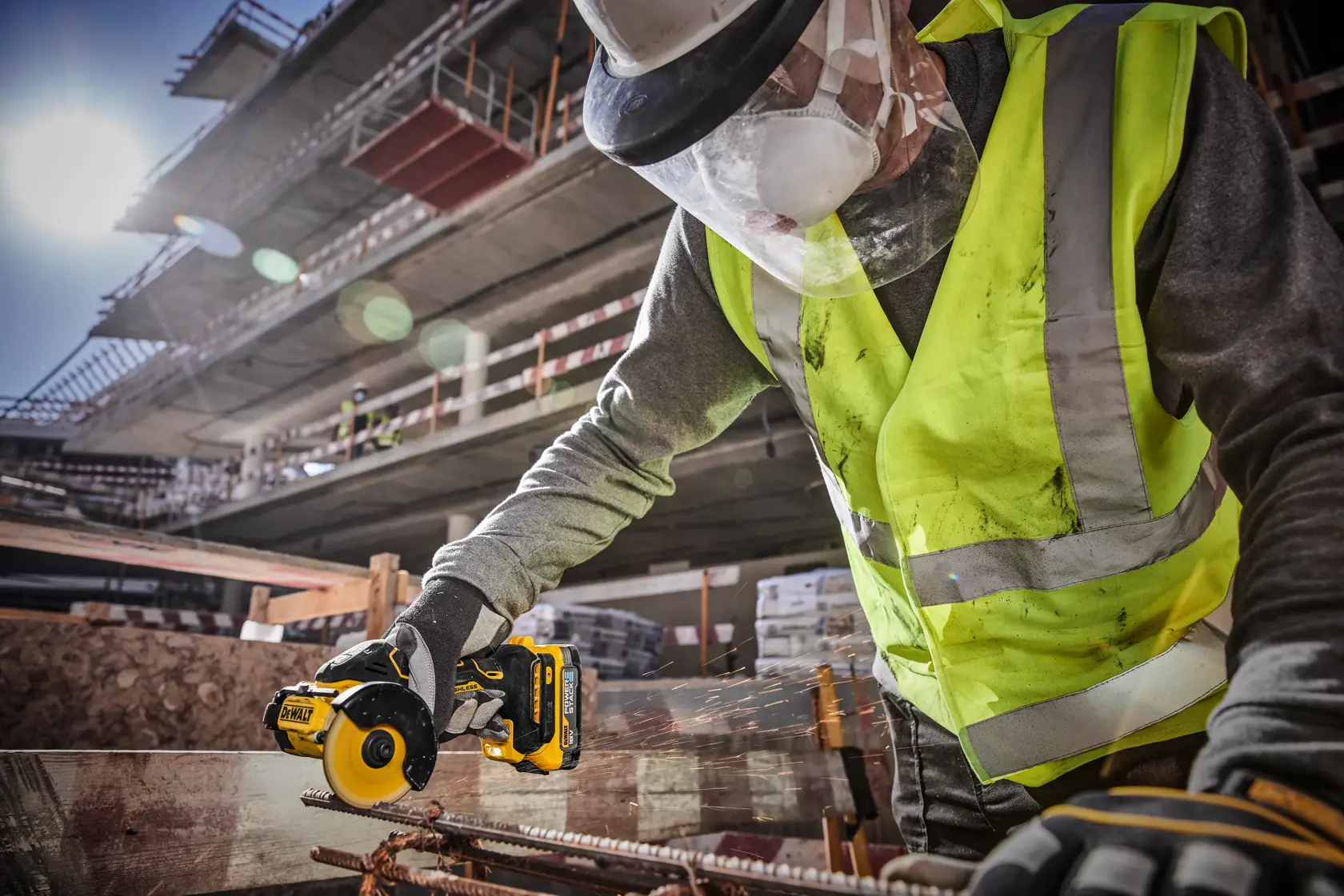 En bygningsarbeider med refleksvest, beskyttelseshansker, ansiktsmaske og hjelm bruker et DEWALT DCS438N batteridrevet verktøy til å kutte metallarmering på en byggeplass med en delvis oppført struktur i bakgrunnen.