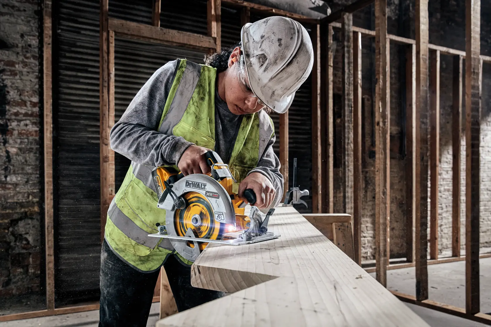 En person med sikkerhetsvest og hjelm bruker en DEWALT DCS573H2T batteridrevet sirkelsag for å kutte en stor treplanke på en uferdig byggeplass.