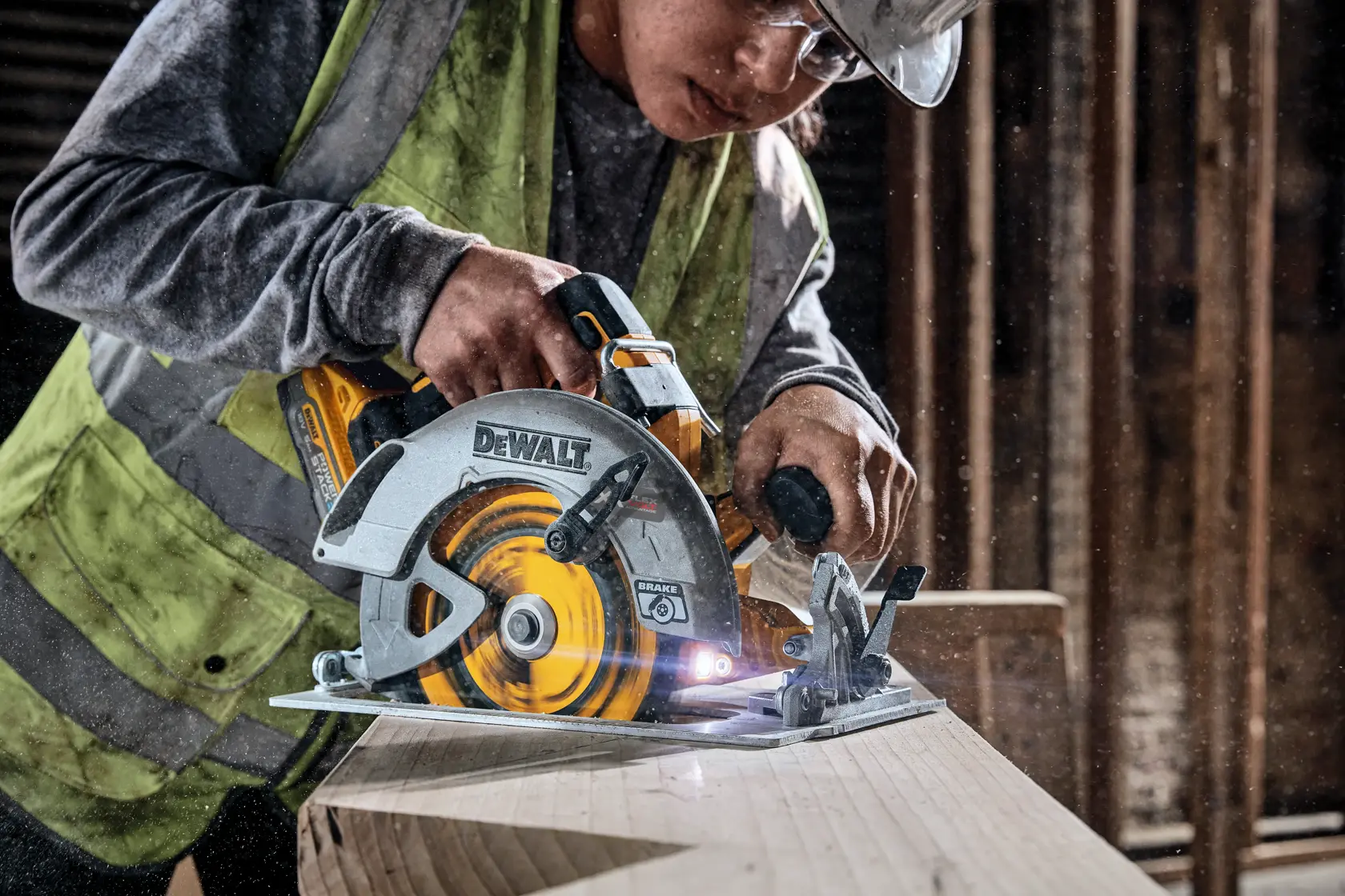 En person med verneutstyr bruker en DEWALT batteridrevet sirkelsag til å skjære en trebjelke på et verksted eller byggeplass.