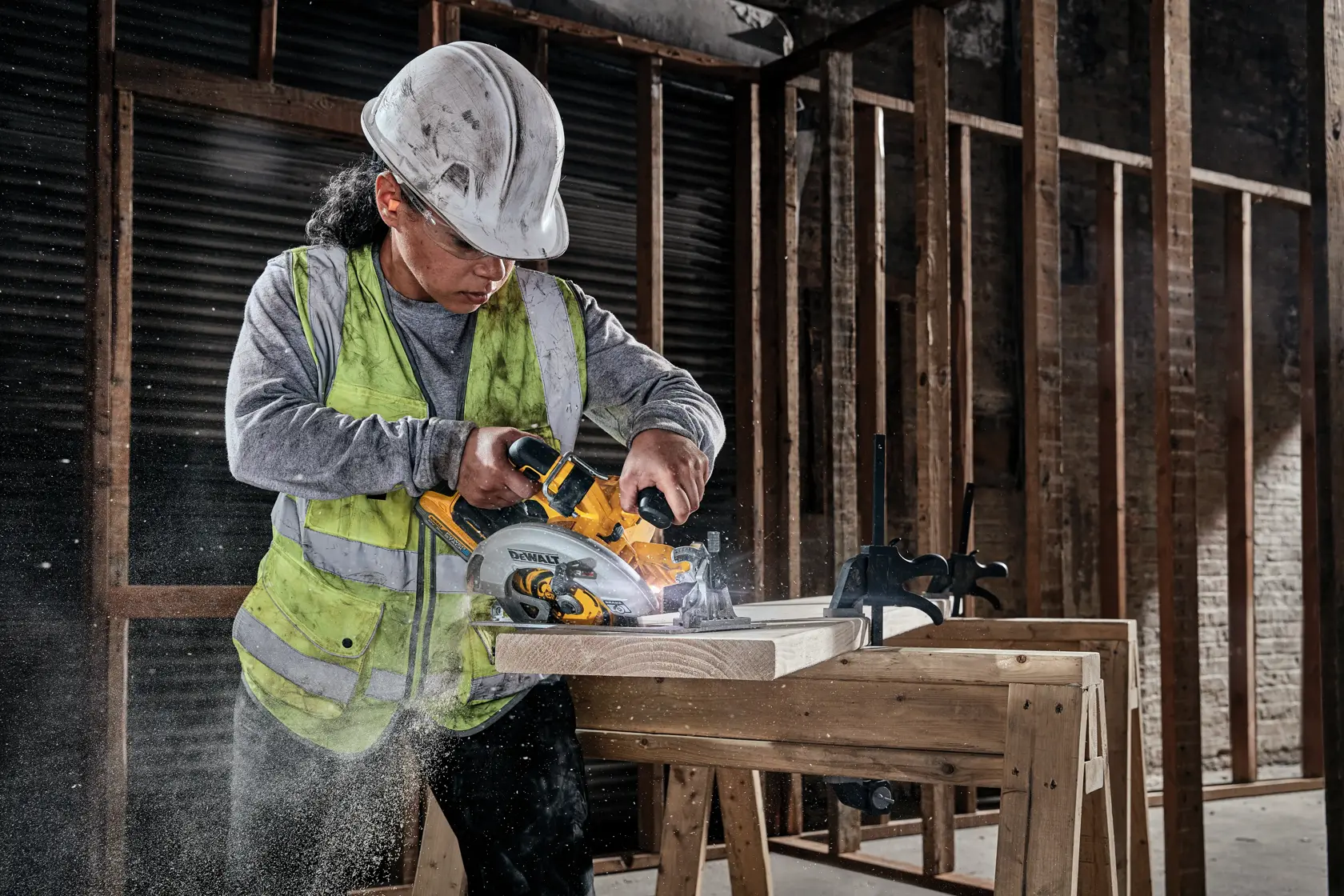 En bygningsarbeider med vernehjelm og refleksvest bruker en trådløs DEWALT sirkelsag til å kutte treplanker på en arbeidsbenk i en uferdig bygning.