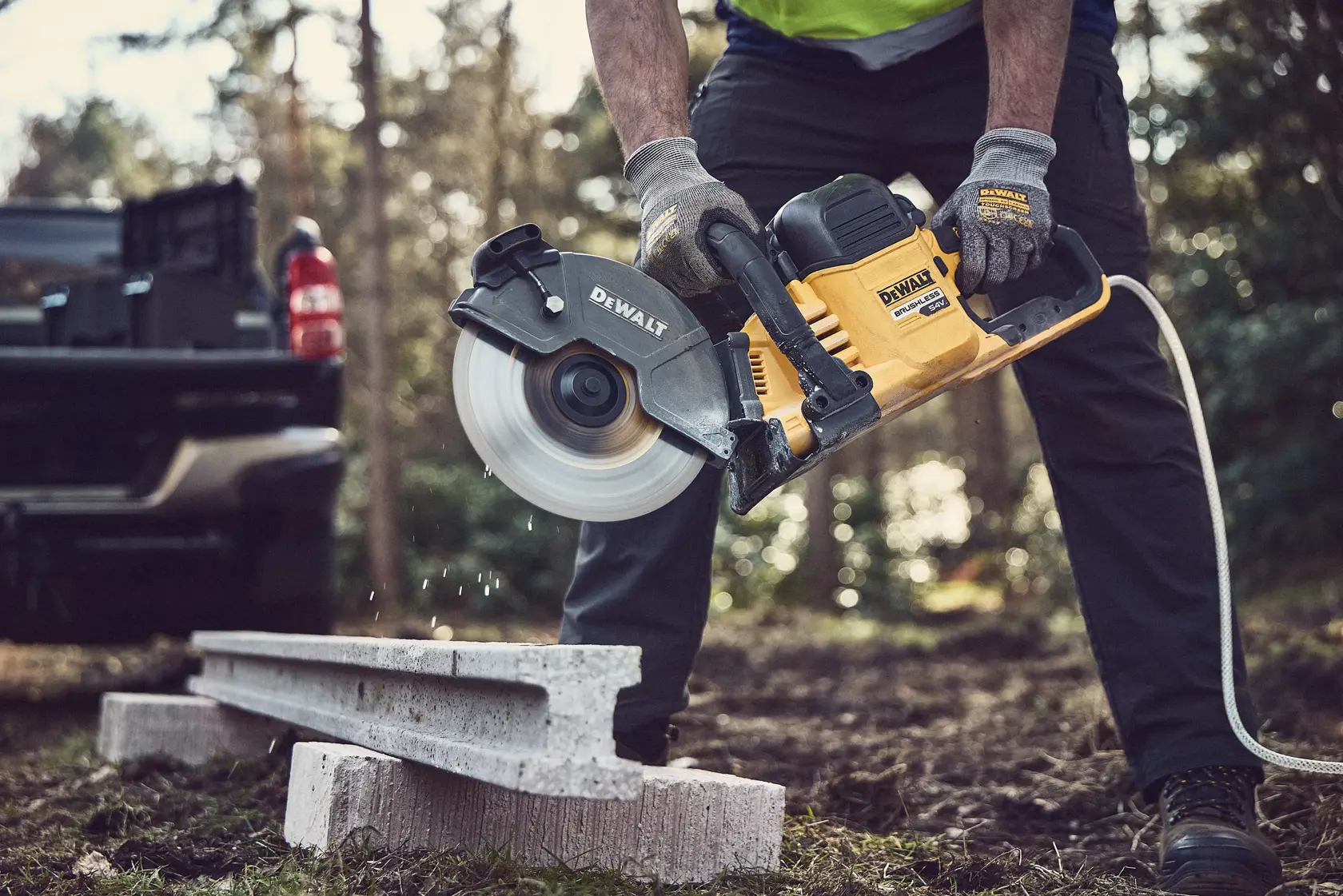 Persona usa una sega DEWALT per tagliare cemento all'aperto. Guanti protettivi e camion visibili.