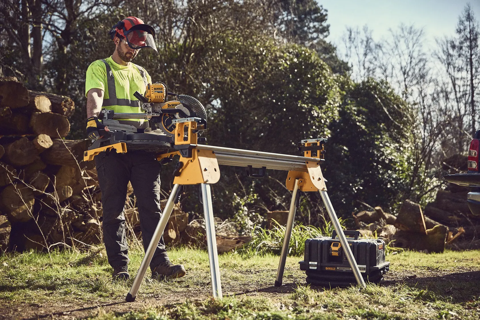 A DEWALT miter saw on a stand outdoors, with a worker using safety gear near logs and a toolbox.