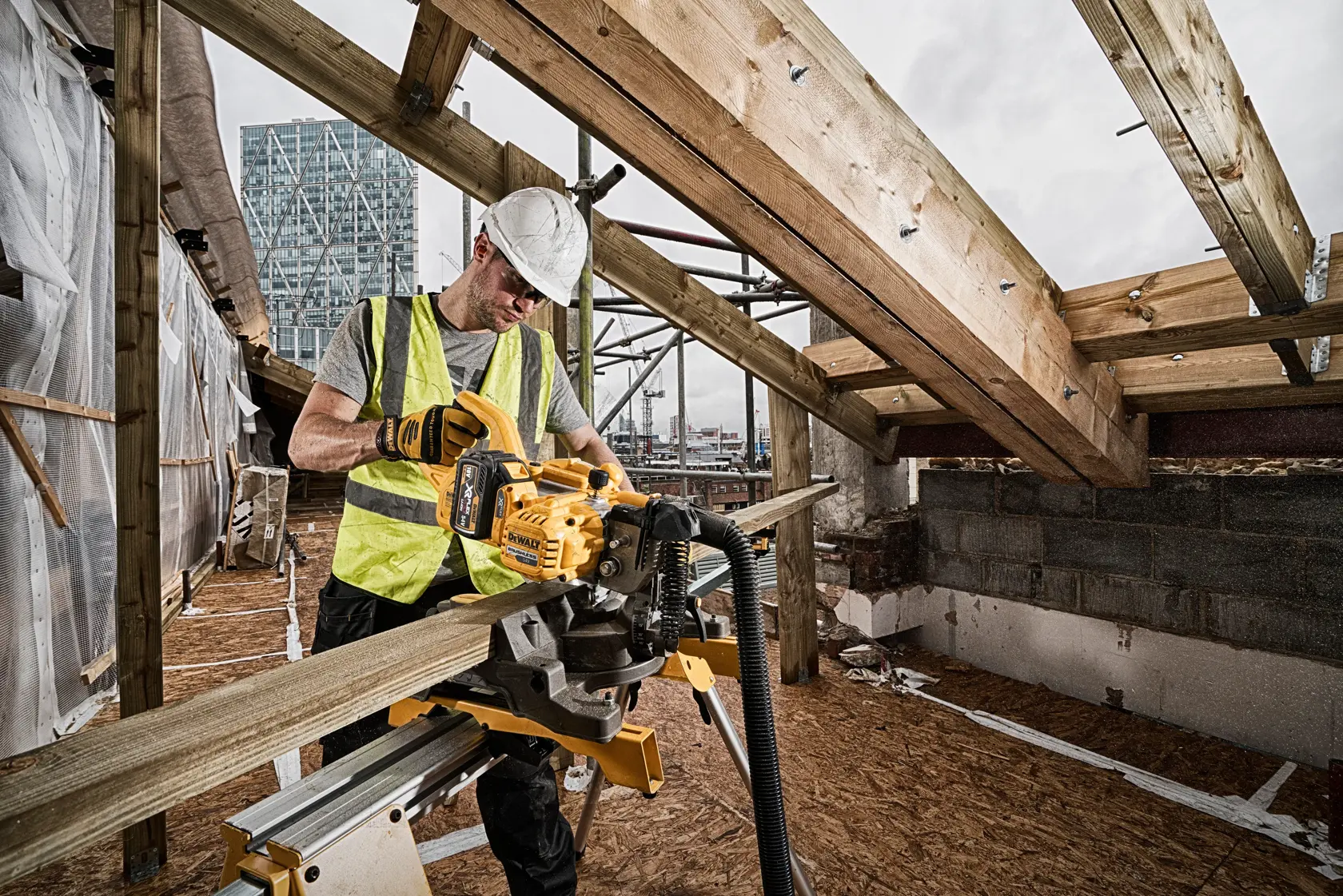 A construction worker wearing a high-visibility vest and a hard hat operates a DEWALT cordless mitre saw (SKU: DCS777N) to cut a wooden beam on a construction site. The surroundings include partially built structures with exposed beams and urban buildings in the background.
