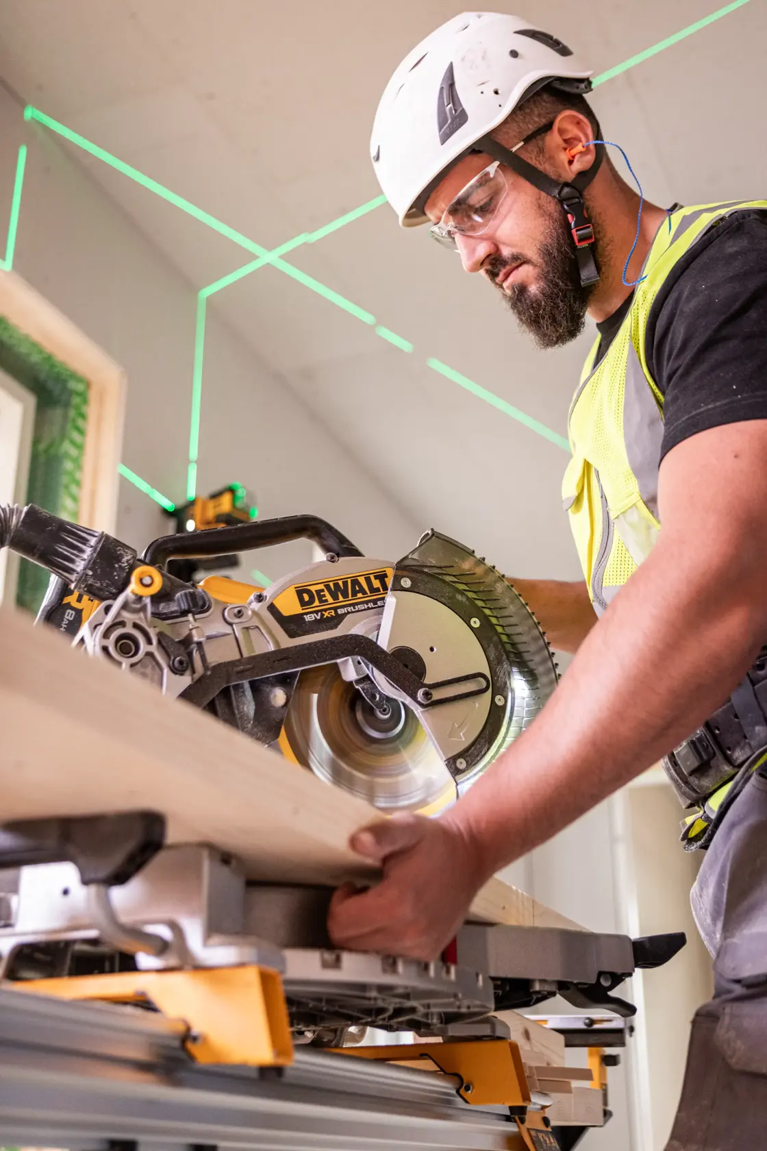 Worker uses DEWALT DCS782N saw to cut wood indoors, wearing safety gear and helmet.