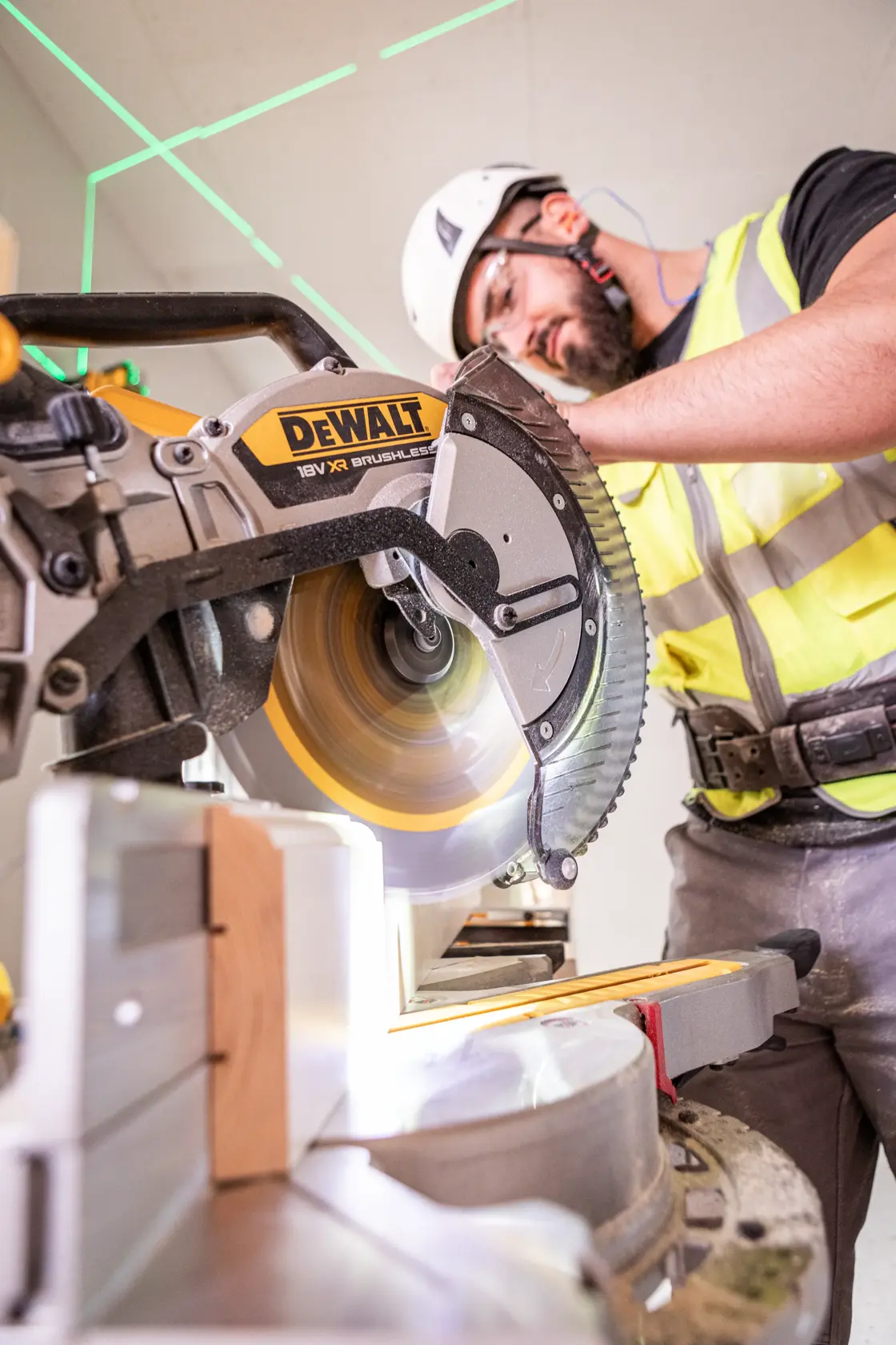 Person using a DEWALT miter saw to cut wood, wearing safety gear at a construction site.
