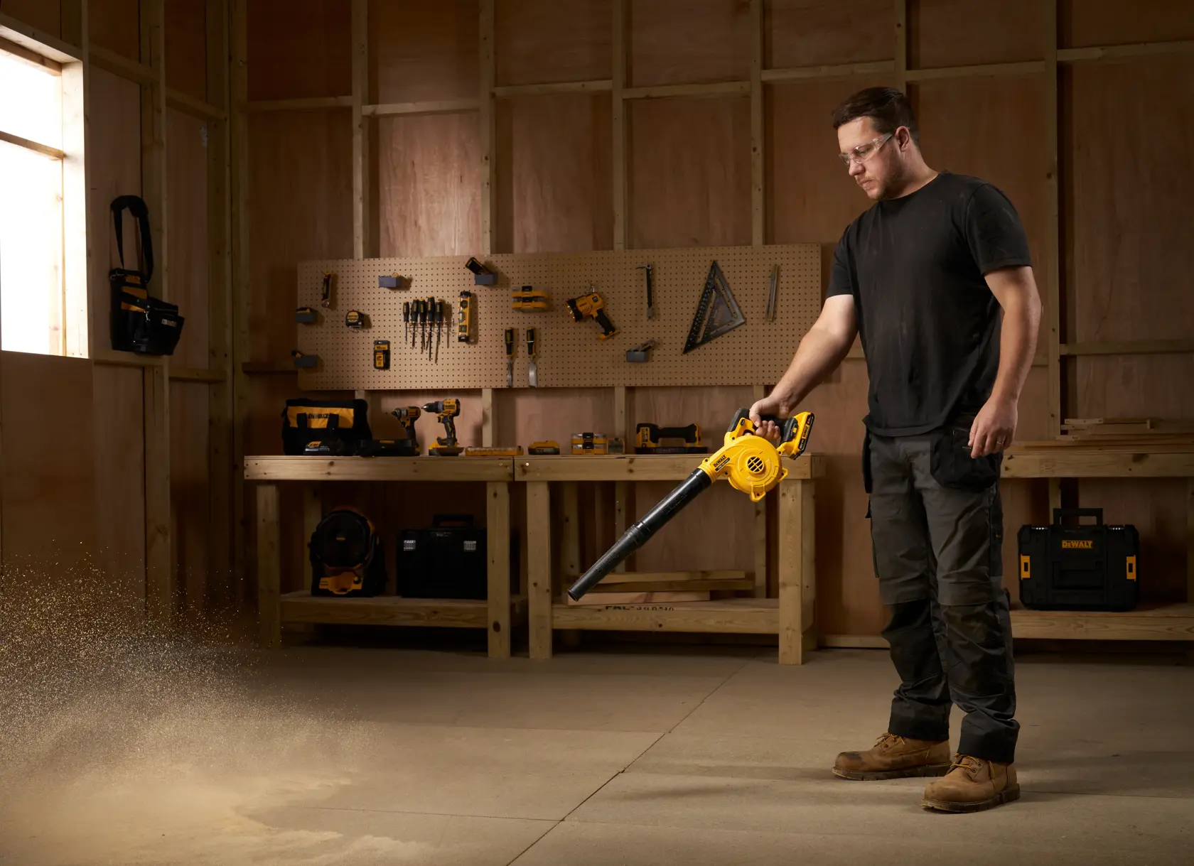 Person uses a DEWALT DCV100 blower to clear dust in a workshop with tools and wooden workbench.