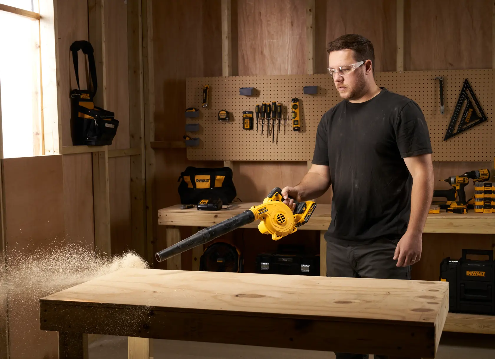 Person using a DEWALT DCV100 blower to clean sawdust off a wooden workbench in a workshop.