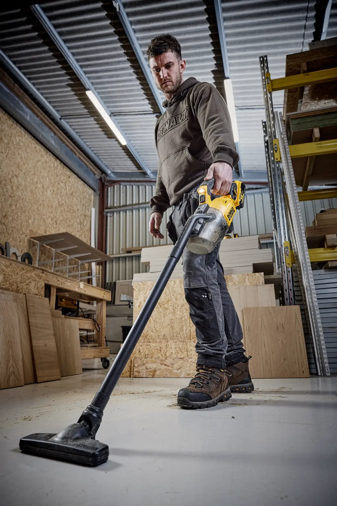 Person using a DEWALT DCV501LN vacuum cleaner on a workshop floor amid wood panels and shelves.