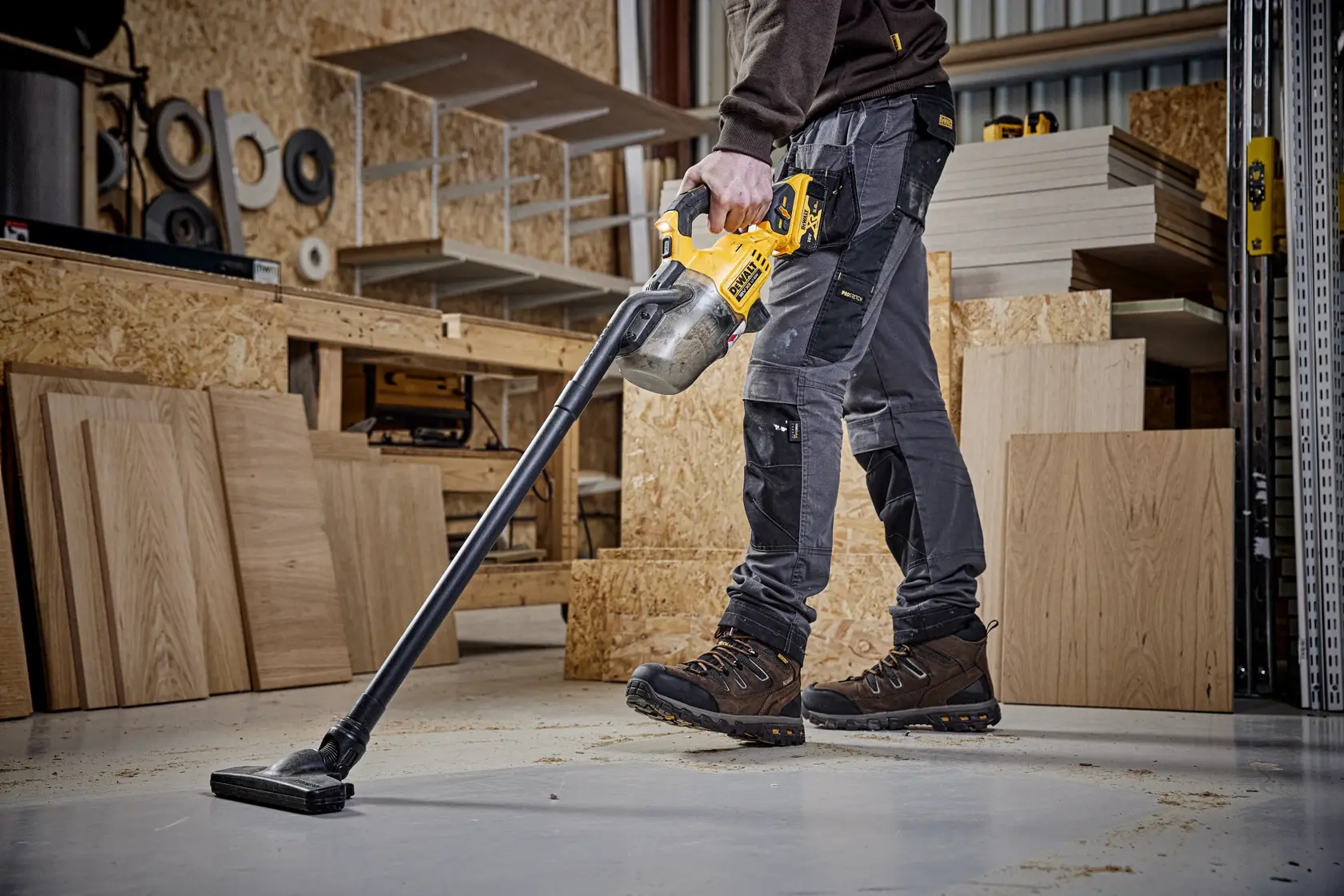 Person using DEWALT cordless vacuum to clean sawdust on workshop floor, surrounded by wood panels.