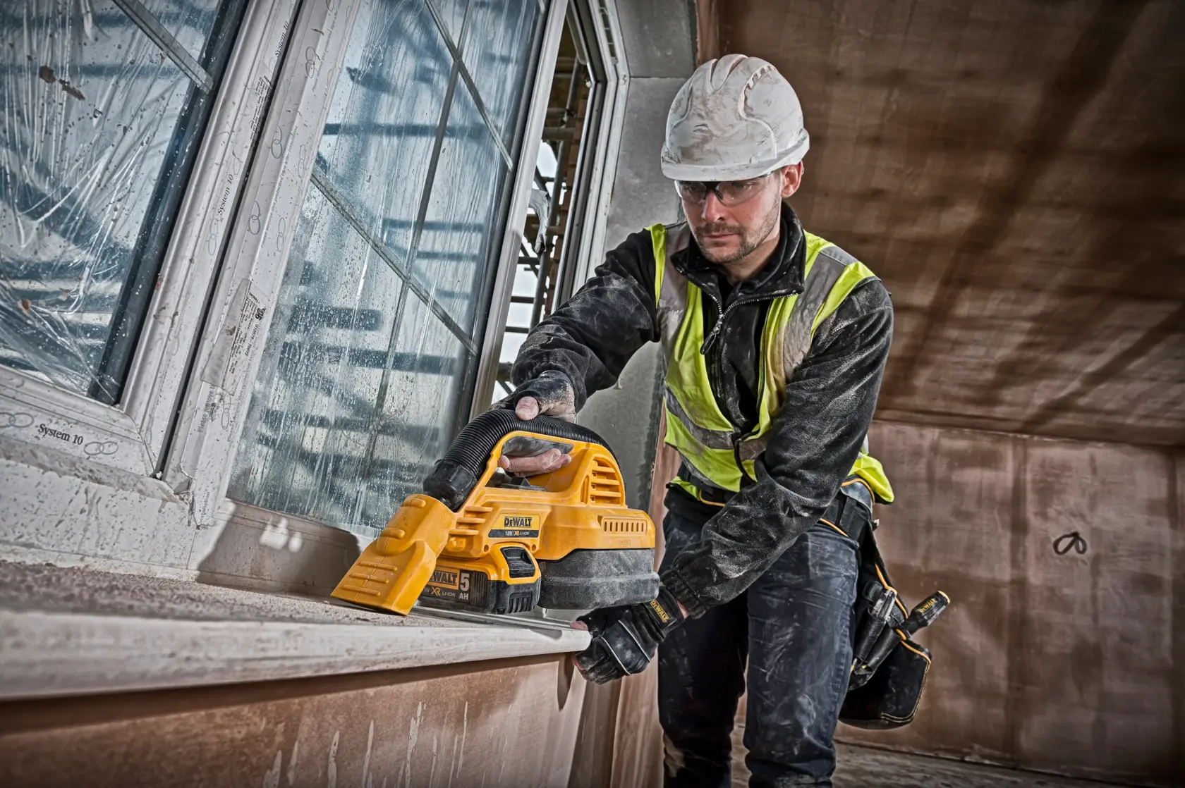 Worker uses DEWALT DCV517N portable vacuum on windowsill at a construction site. Safety gear and high-vis vest.